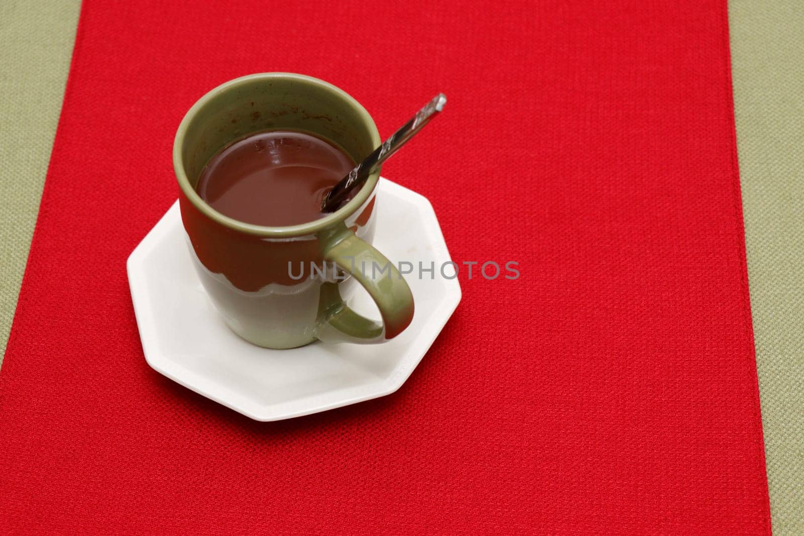 A cup of hot cocoa mix with spoon on a white saucer placed on a red place mat on a green tablecloth. Warm hot chocolate mix in a cup with a metal spoon on a red and green textile tabletop background.
