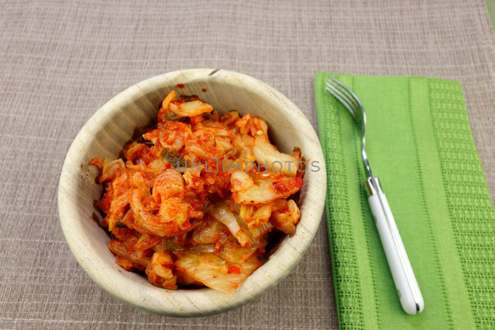 Fermented meal of napa cabbage, radish and seasonings kimchi recipe ready to eat from a round wood bowl. Ready to eat kimchi recipe in a wood bowl on a brown place mat near a fork and green napkin..