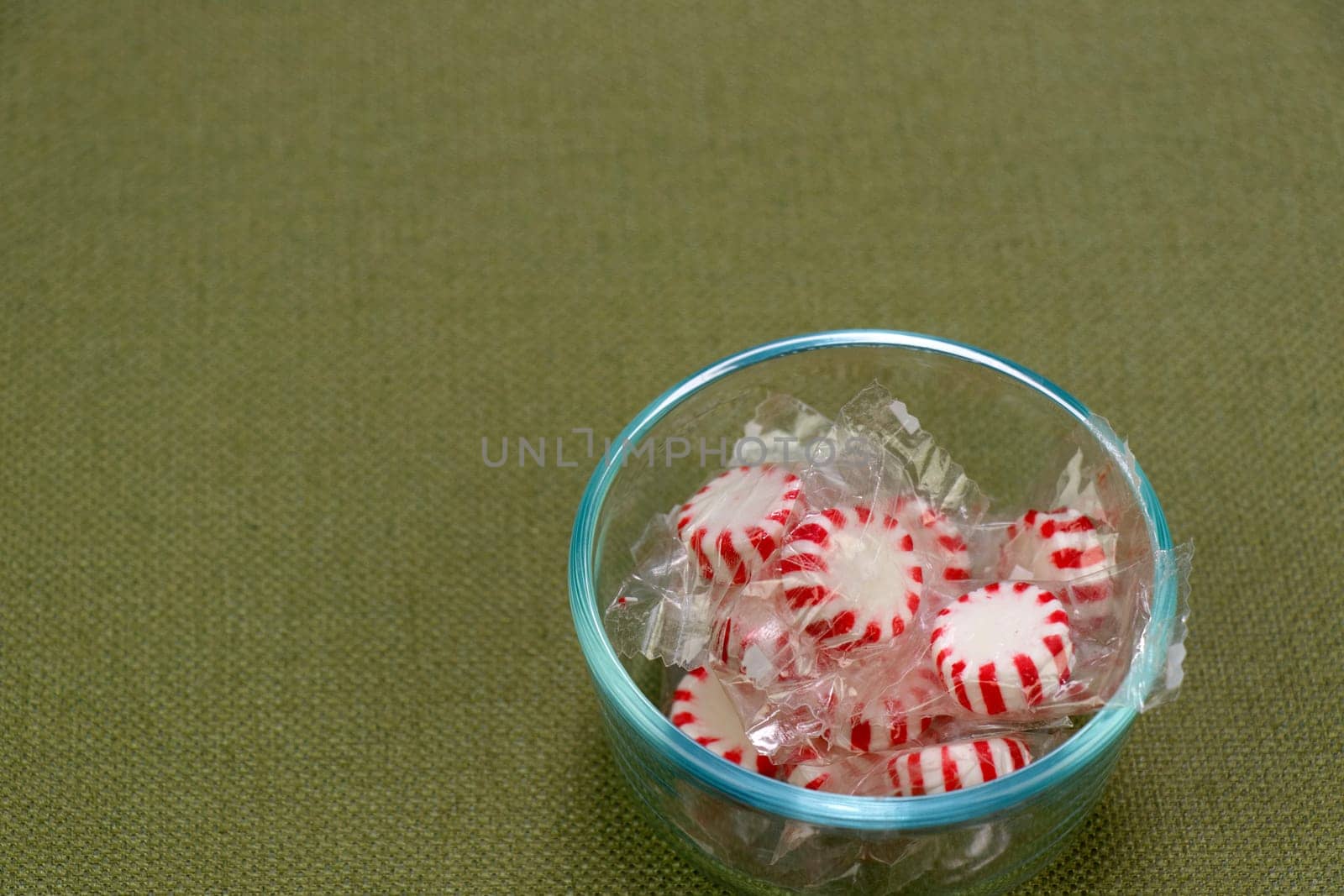 Clear plastic wrapped red and white peppermint hard candies in a small clear glass bowl on a green tablecloth. Holiday white and red peppermint hard candy discs in a small bowl on a green tablecloth.