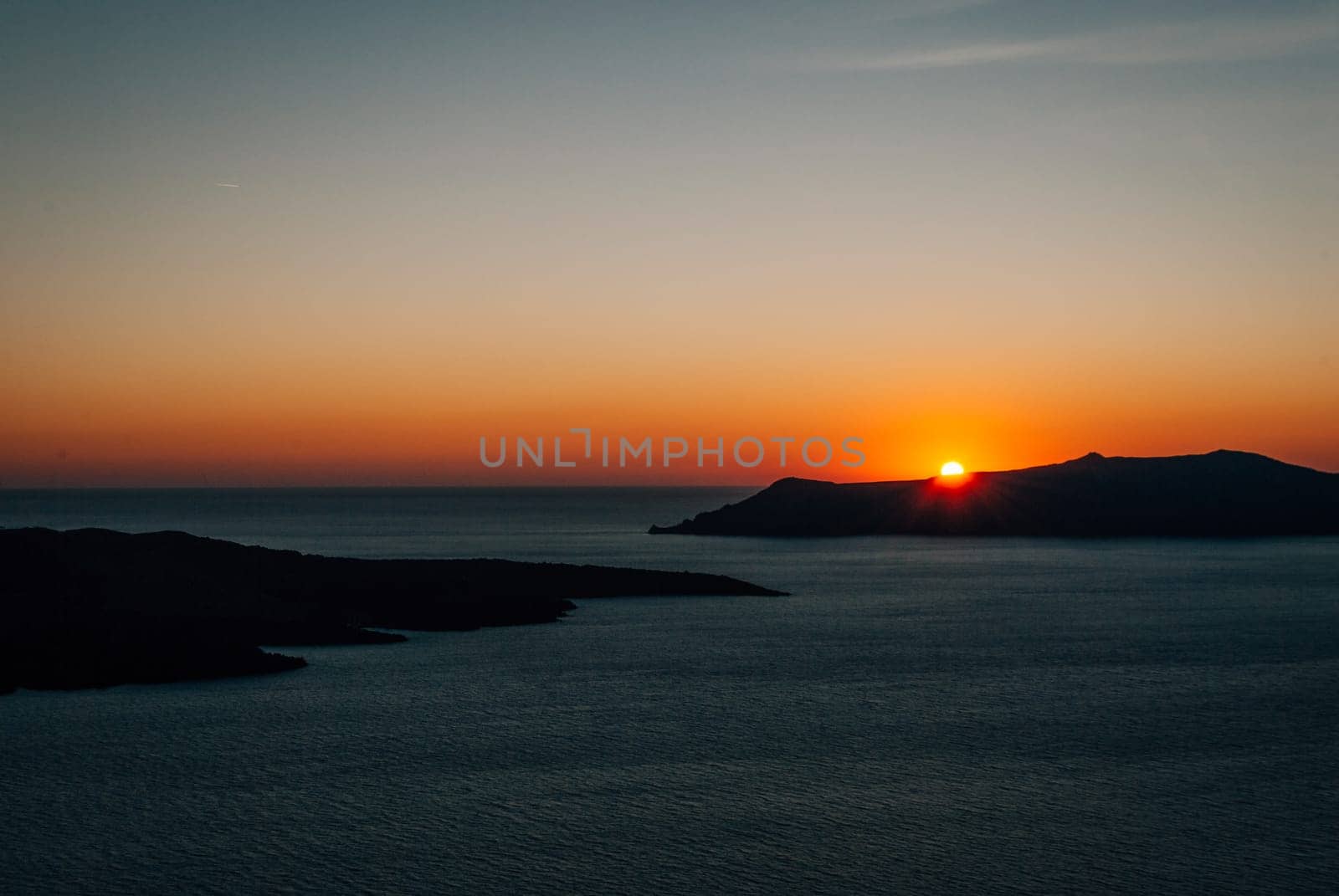 The sun sets behind an island in the sea, Santorini, Greece.