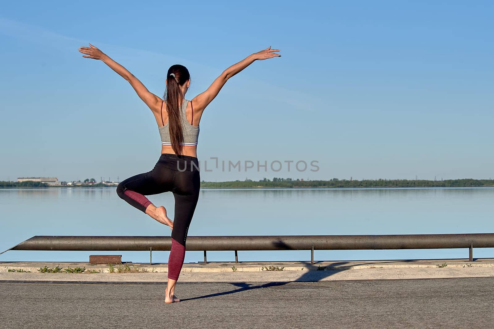 A young woman does a warm-up on the pier in the early morning, against the background of the river. High quality photo