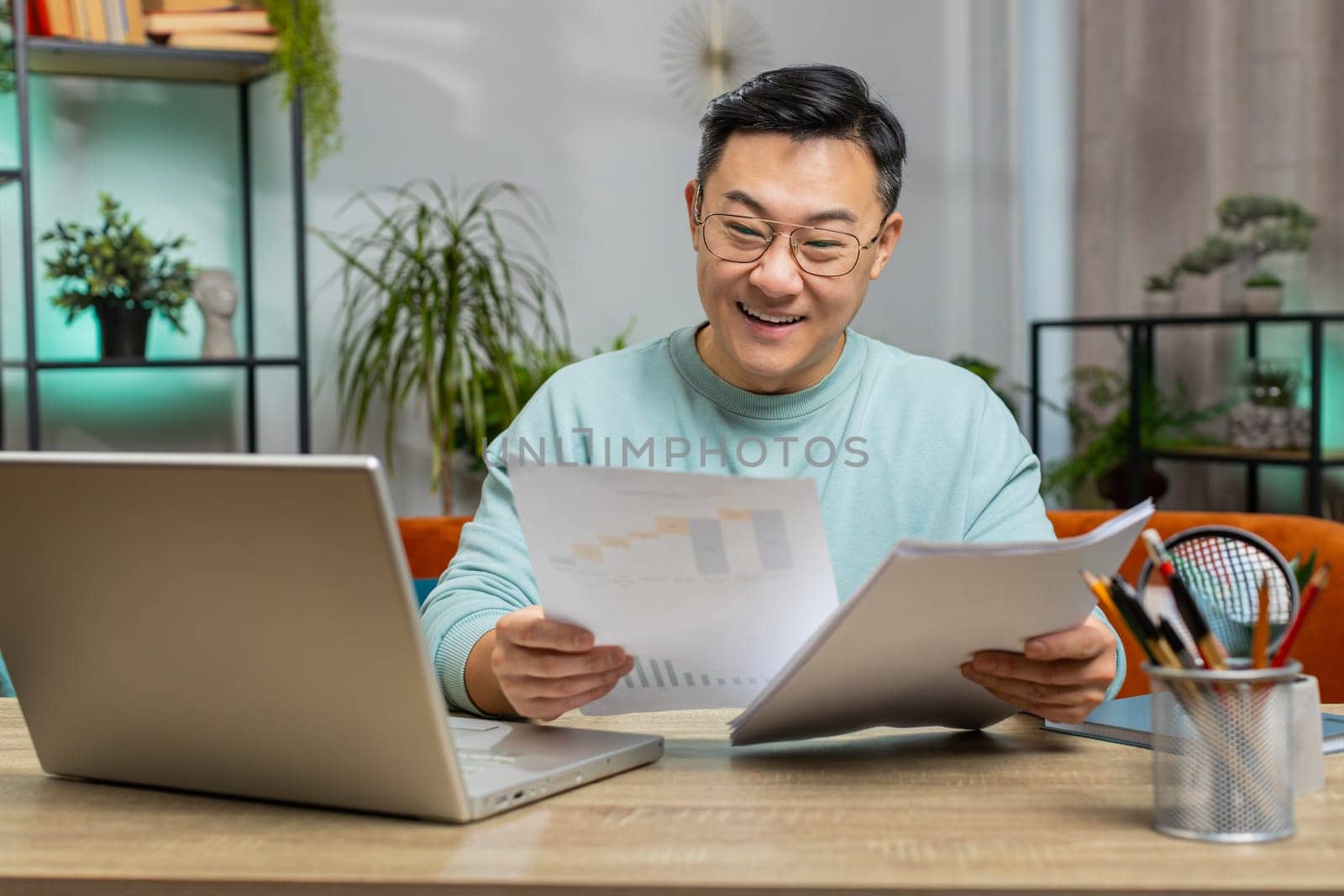 Happy Asian man freelancer work with documents at home analyzing information read business papers prepare financial report. Guy winner shocked by victory, good success news sits on couch at table desk