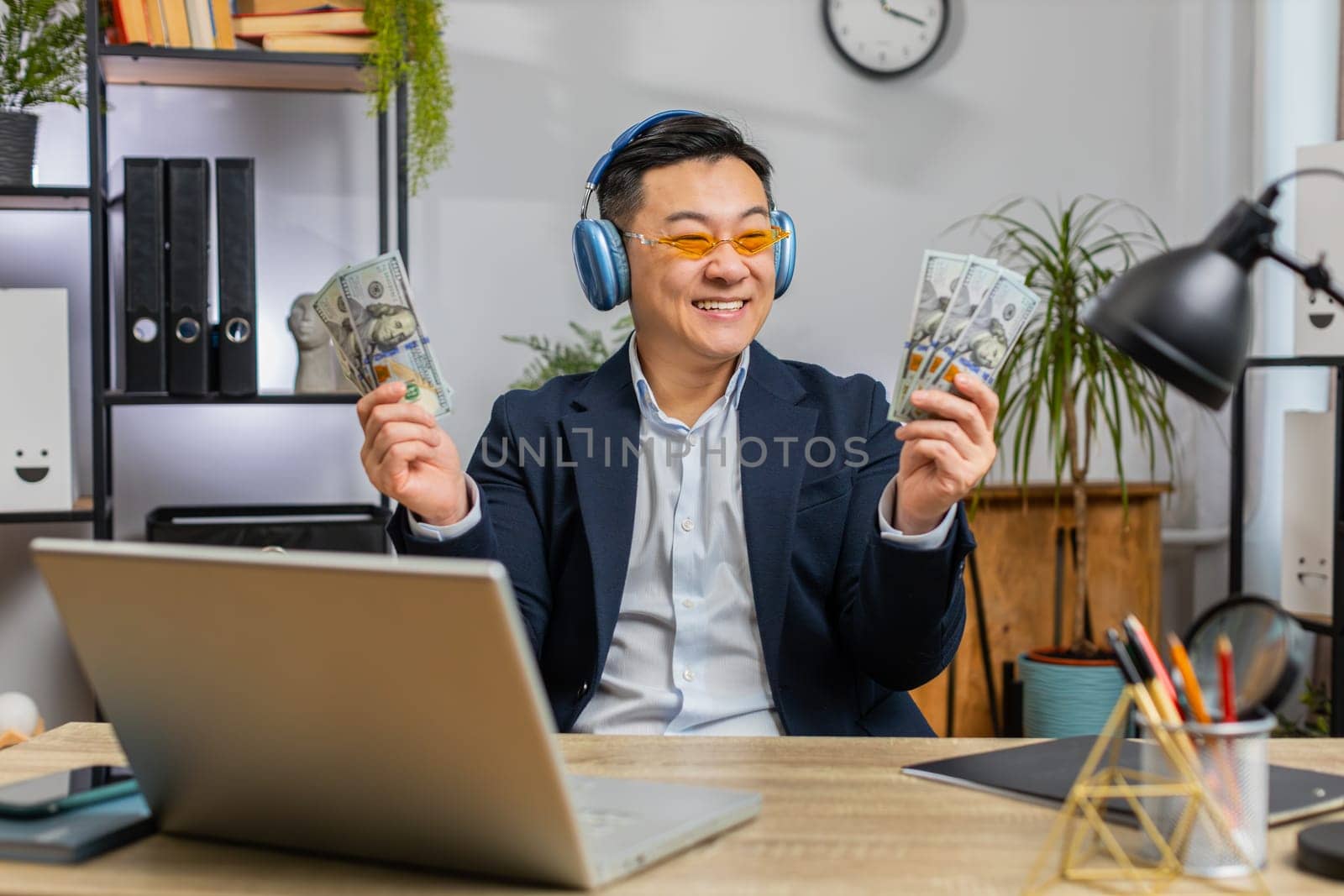 Asian Chinese businessman holding fan of cash money dollar banknotes celebrate, dance, success career, lottery jackpot game winner, big income, wealth at office workplace desk. Remote distant working