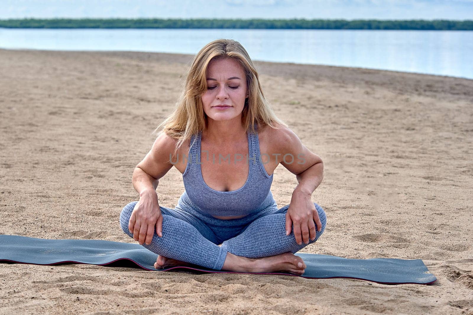 A woman does yoga on a sandy beach against the background of a river. High quality photo