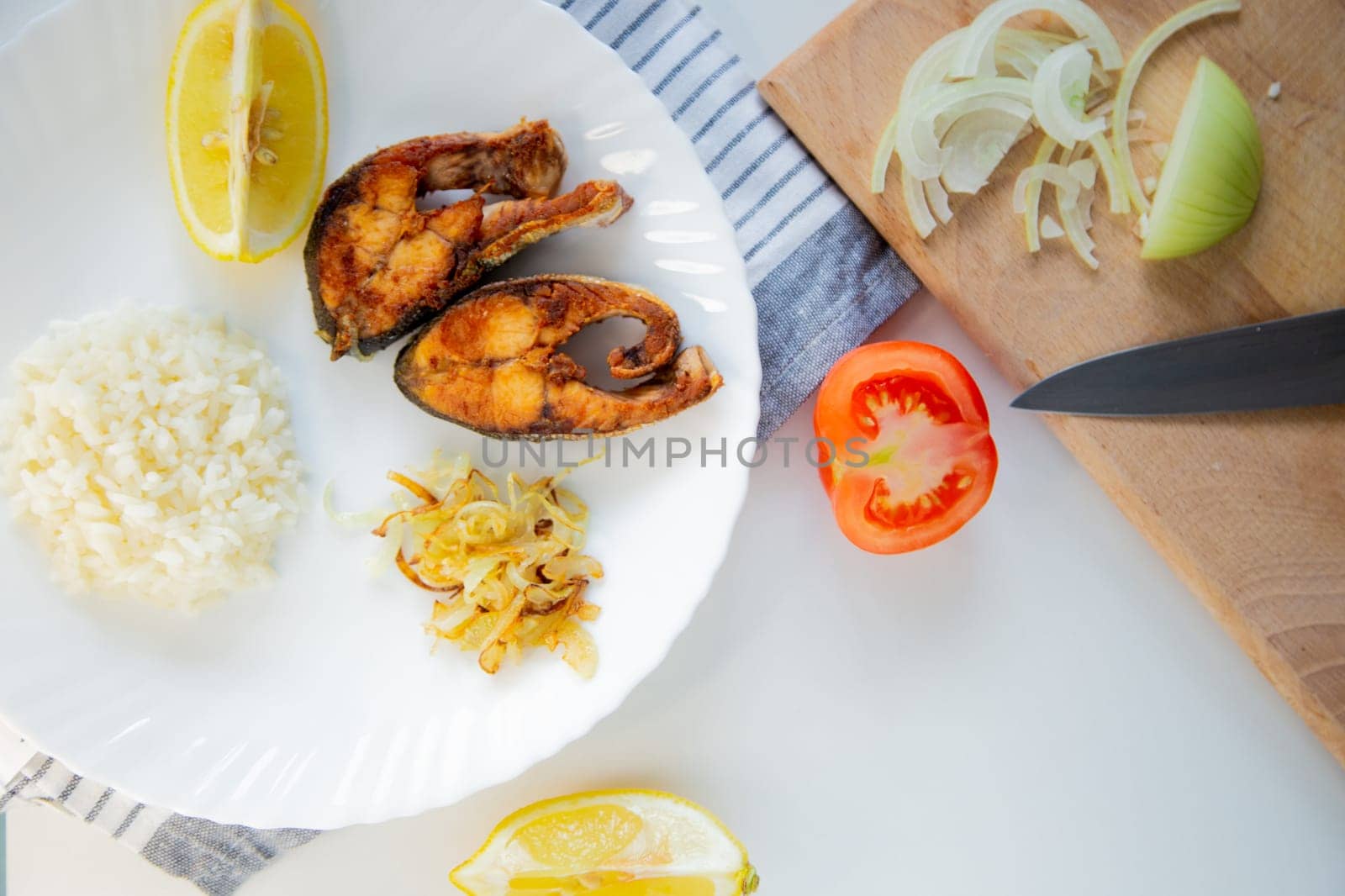 Fried fish with onions and rice is on a plate, on a waffle towel, next to it is a cutting board with chopped onions, lemon and tomato on a white background. High quality photo
