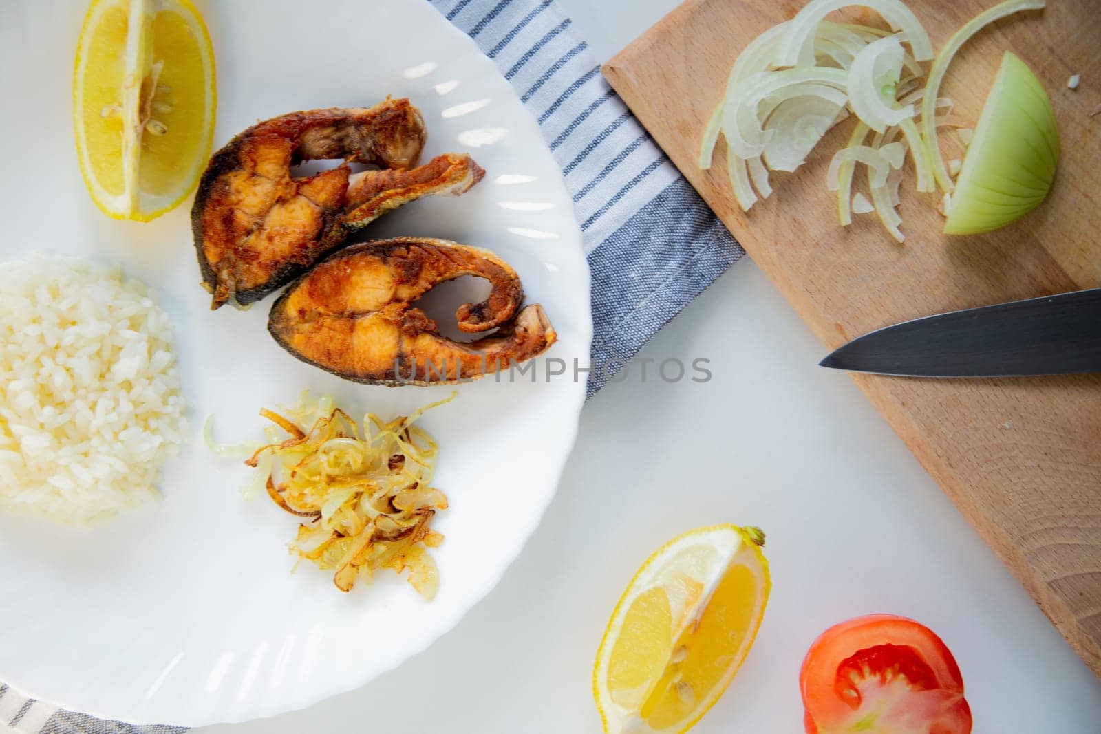 Fried fish with onions and rice is on a plate, on a waffle towel, next to it is a cutting board with chopped onions, lemon and tomato on a white background. High quality photo