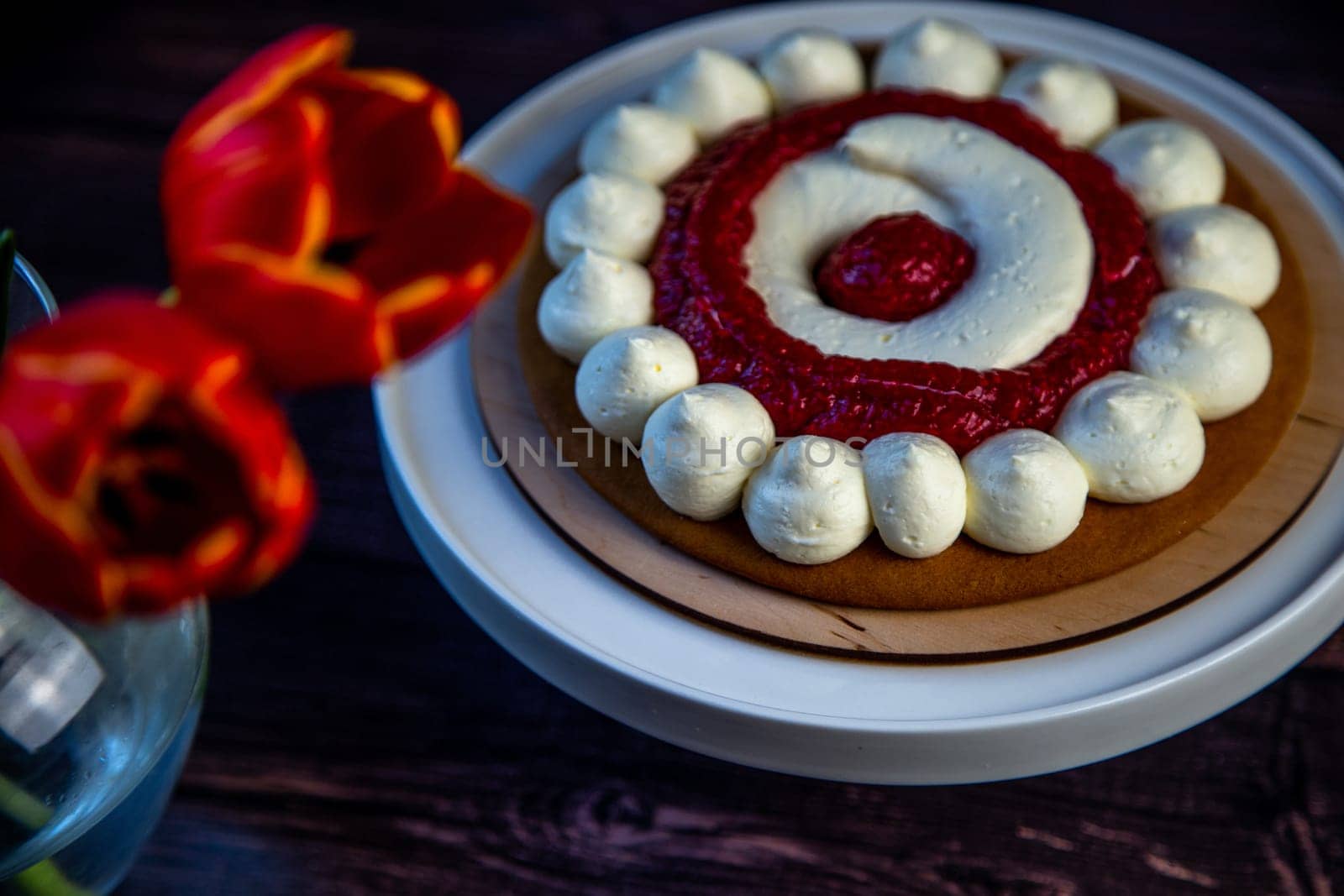 The baked cake cake lies on a wooden substrate and on a white stand, the cake is decorated with white cream and raspberry jam, in a circle, circles alternate, a bouquet of red tulips on a dark background is visible nearby. High quality photo