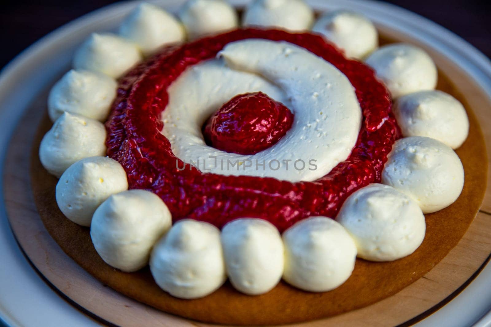 The baked cake cake lies on a wooden substrate and on a white stand, the cake is decorated with white cream and raspberry jam, in a circle, circles alternate on a dark background. High quality photo