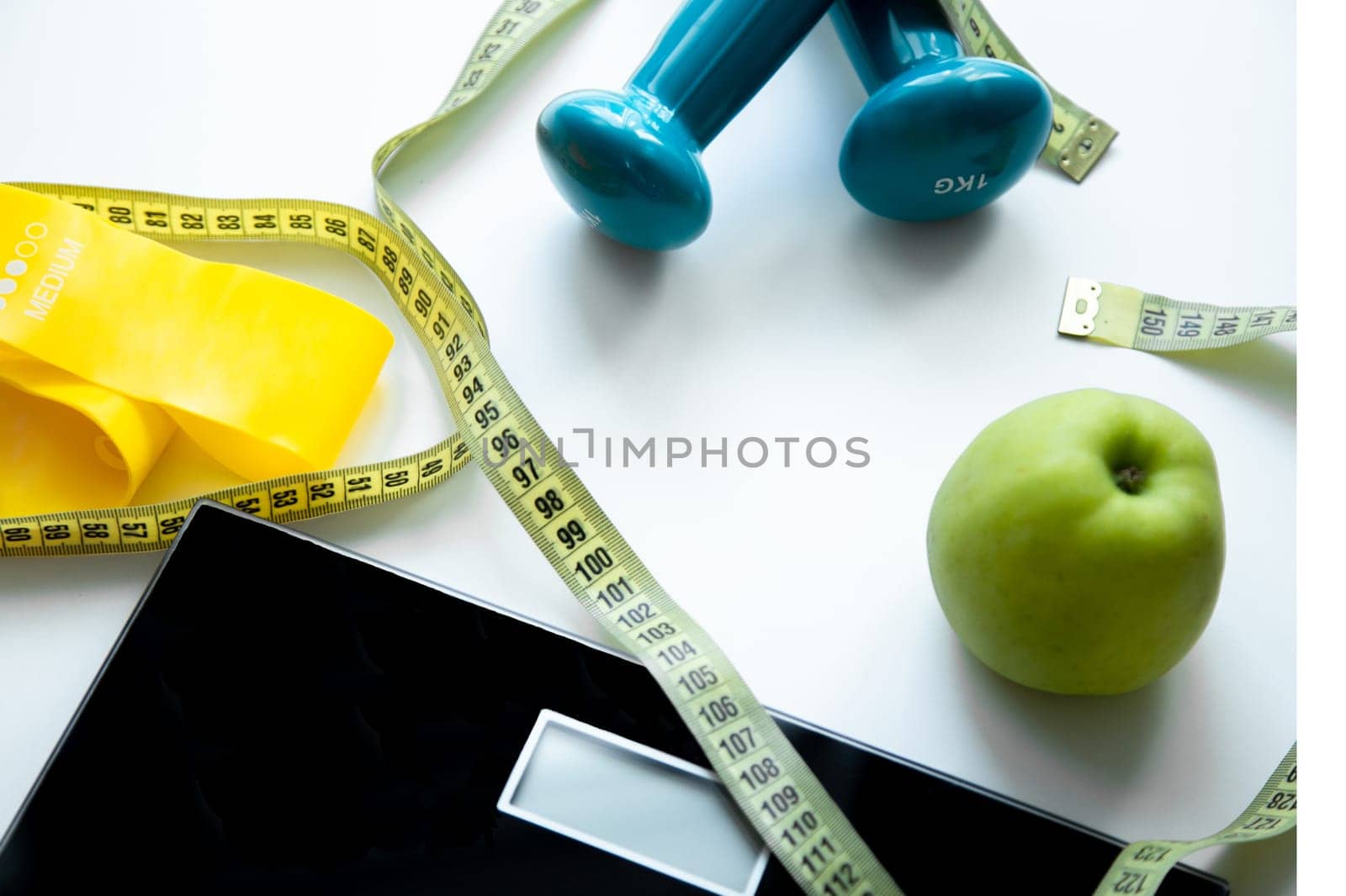 An apple, a scale, a fitness elastic band and dumbbells lie on a white background, they are entwined with a measuring tape. High quality photo
