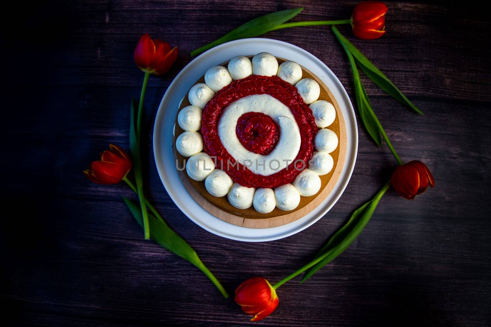 Honey sponge cake with a decoration of white air cream and layers of red raspberries stands on a white platter on a dark wooden background, tulips lie around in a circle. High quality photo