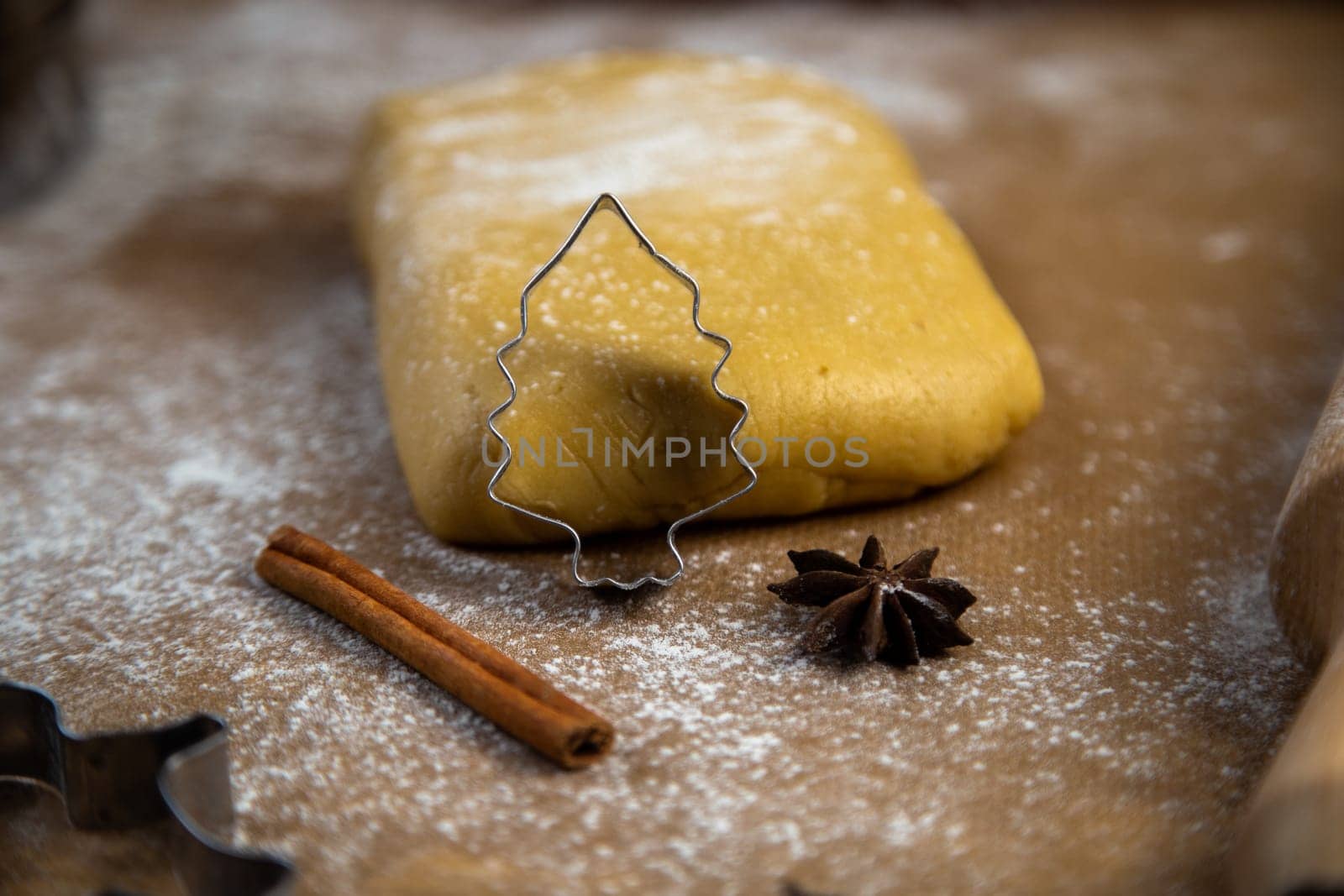 A cookie cutter in the form of a Christmas tree is leaning on the dough, cinnamon and cloves are lying next to it, flour, other cookie cutters and a rolling pin for dough are visible in the background, and everything is in flour. High quality photo