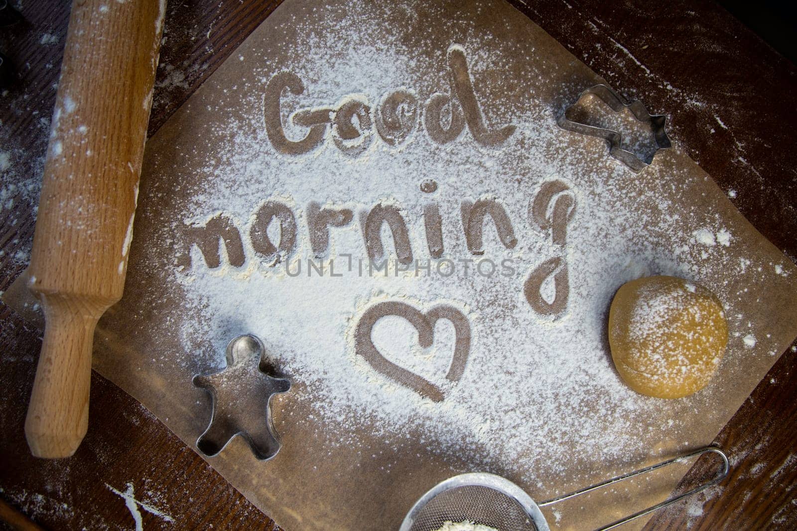 On the baking paper on the flour is written Good morning and heart, against the background of dough, rolling pins and cookie cutters. High quality photo