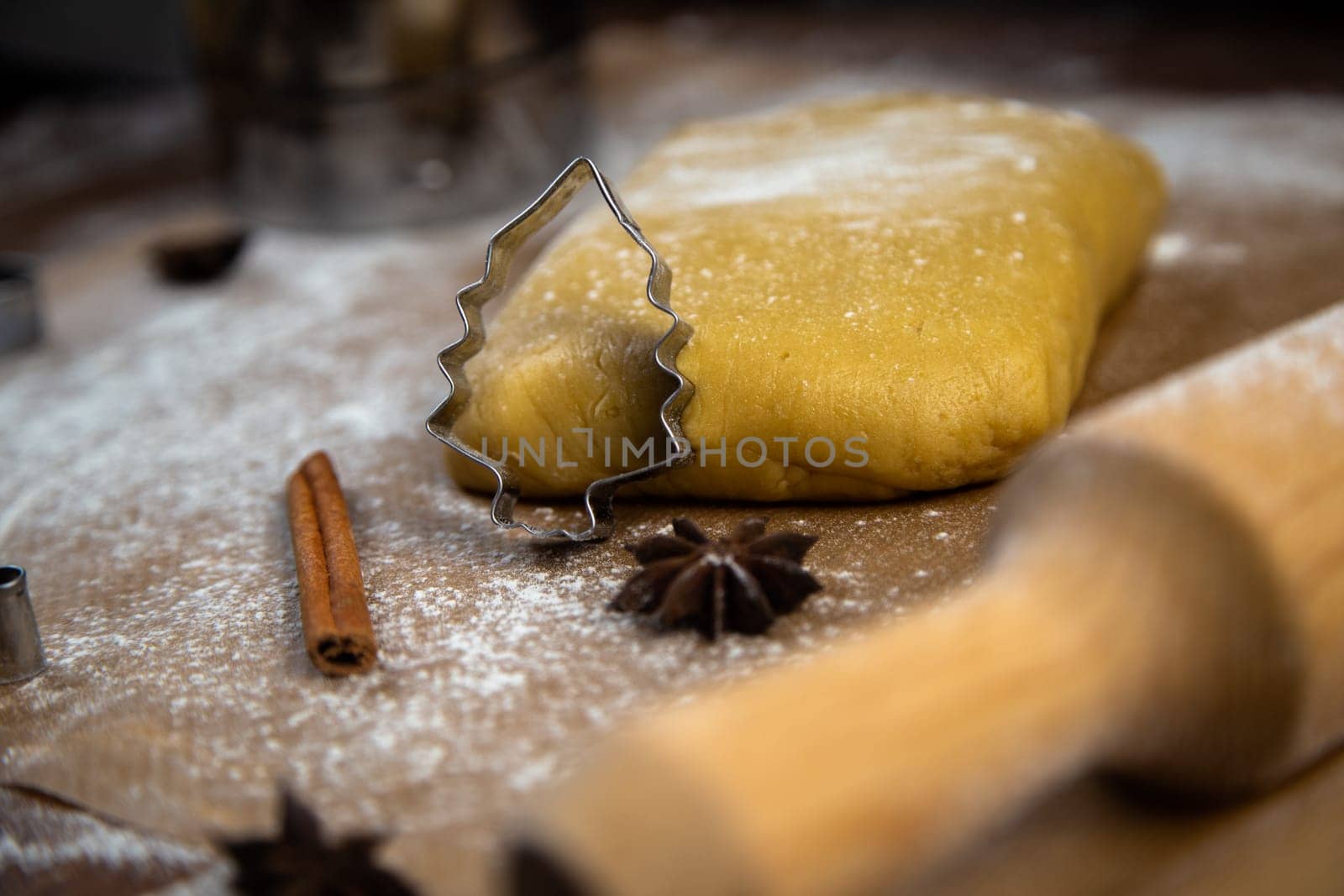 A cookie cutter in the form of a Christmas tree is leaning on the dough, cinnamon and cloves are lying next to it, flour, other cookie cutters and a rolling pin for dough are visible in the background, and everything is in flour. High quality photo
