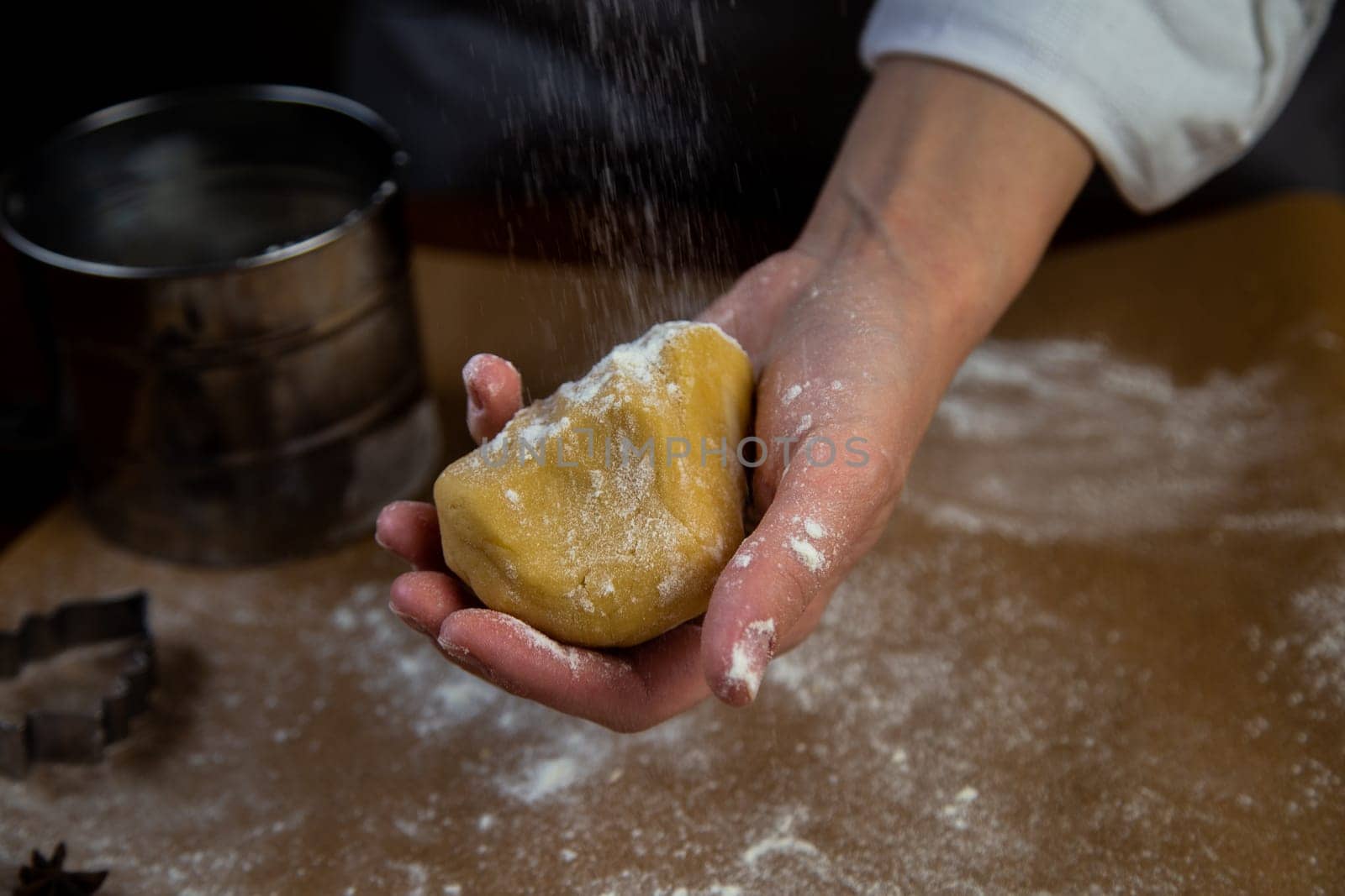In the foreground, there is a piece of dough in his hand, which is sprinkled with flour on top, the background is blurred, everything is in dark colors. High quality photo