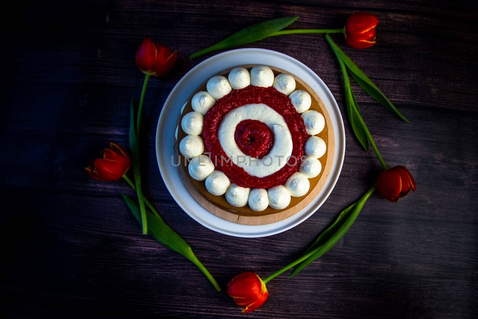 Honey sponge cake with a decoration of white air cream and layers of red raspberries stands on a white platter on a dark wooden background, tulips lie around in a circle. High quality photo