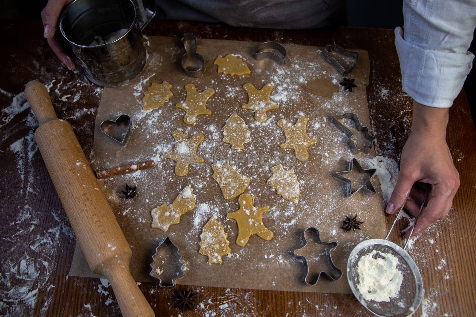 There are ready-made cookies of various shapes on the baking parchment, a rolling pin, cookie cutters and a sieve with flour are lying nearby, flour is scattered everywhere, the hands of the pastry chef are visible. High quality photo