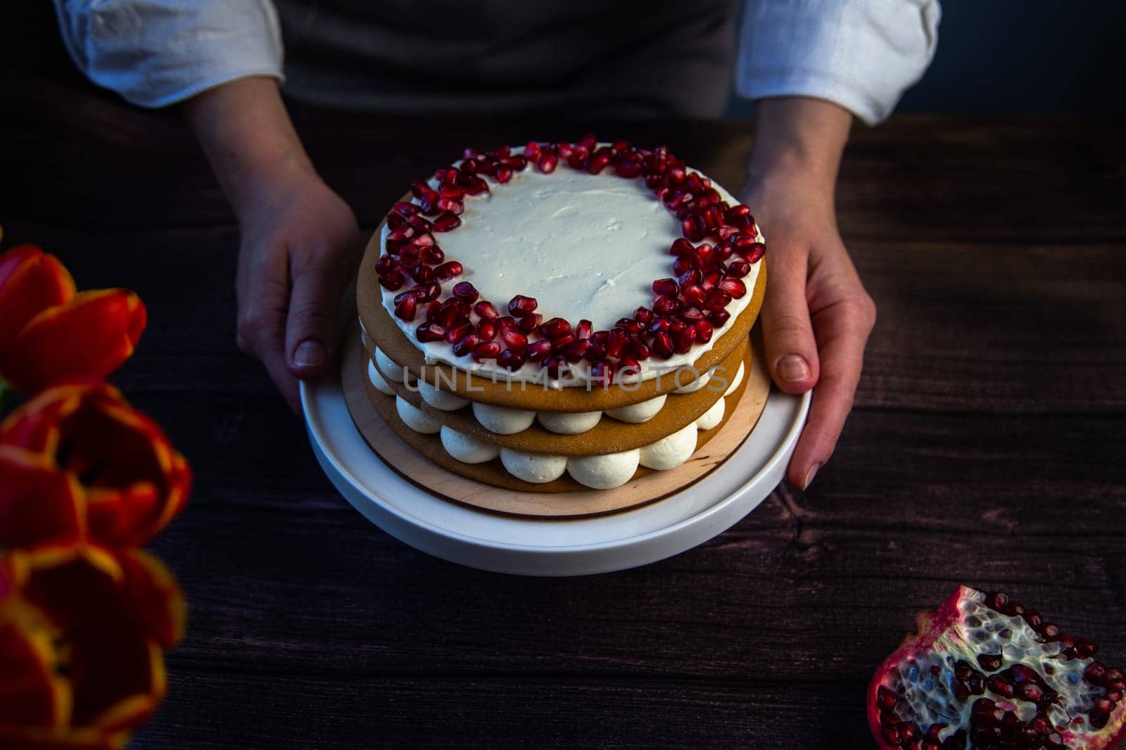 The cake stands on a white stand, decorated with cream and pomegranate on top, it is held by the hands of a pastry chef, there is a pomegranate next to it and there are flowers on a dark background. High quality photo
