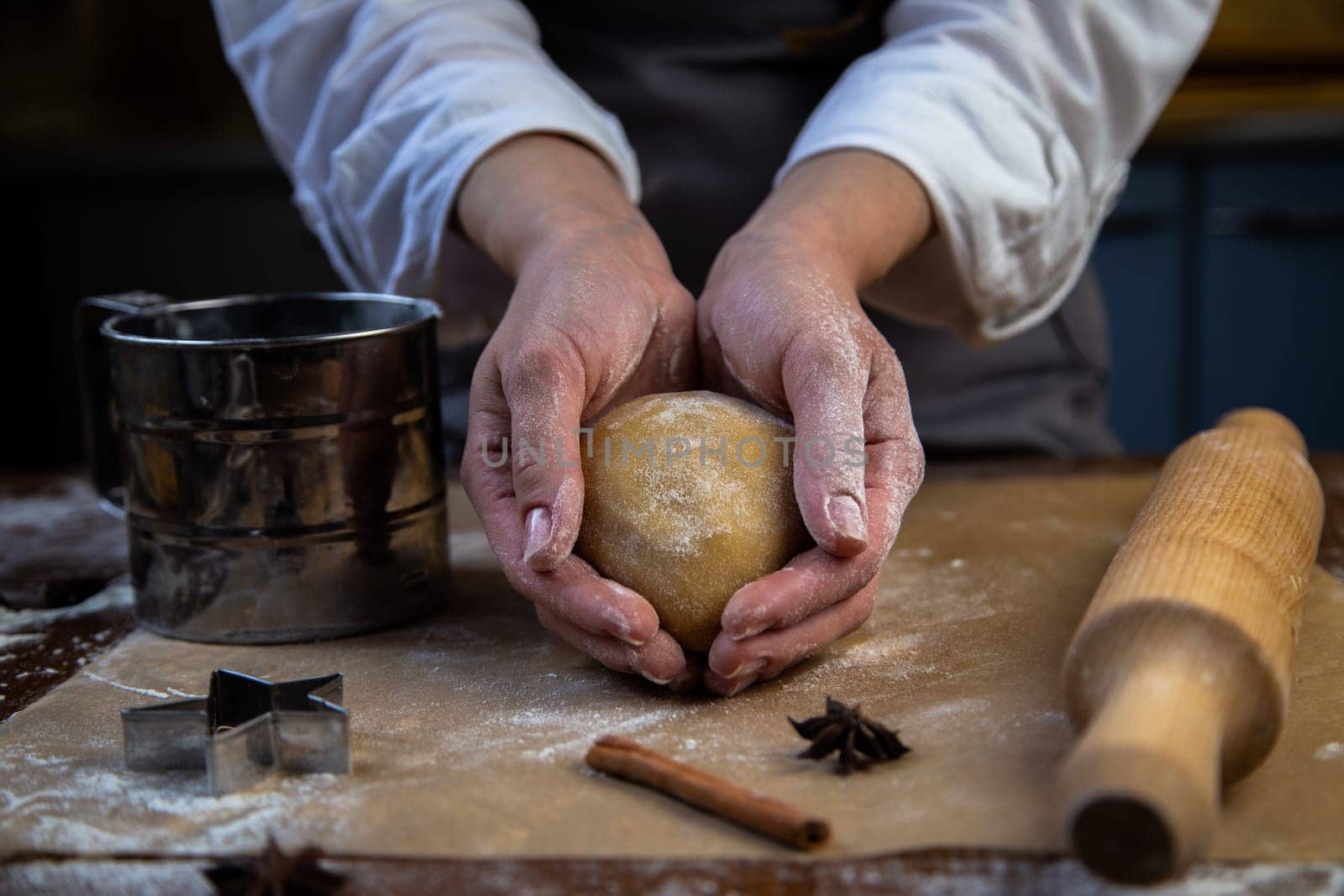 In the hands of a pastry chef, a round piece of dough, all in flour, a rolling pin, a mold with flour, cloves and cinnamon, all in dark colors, lies next to it. High quality photo