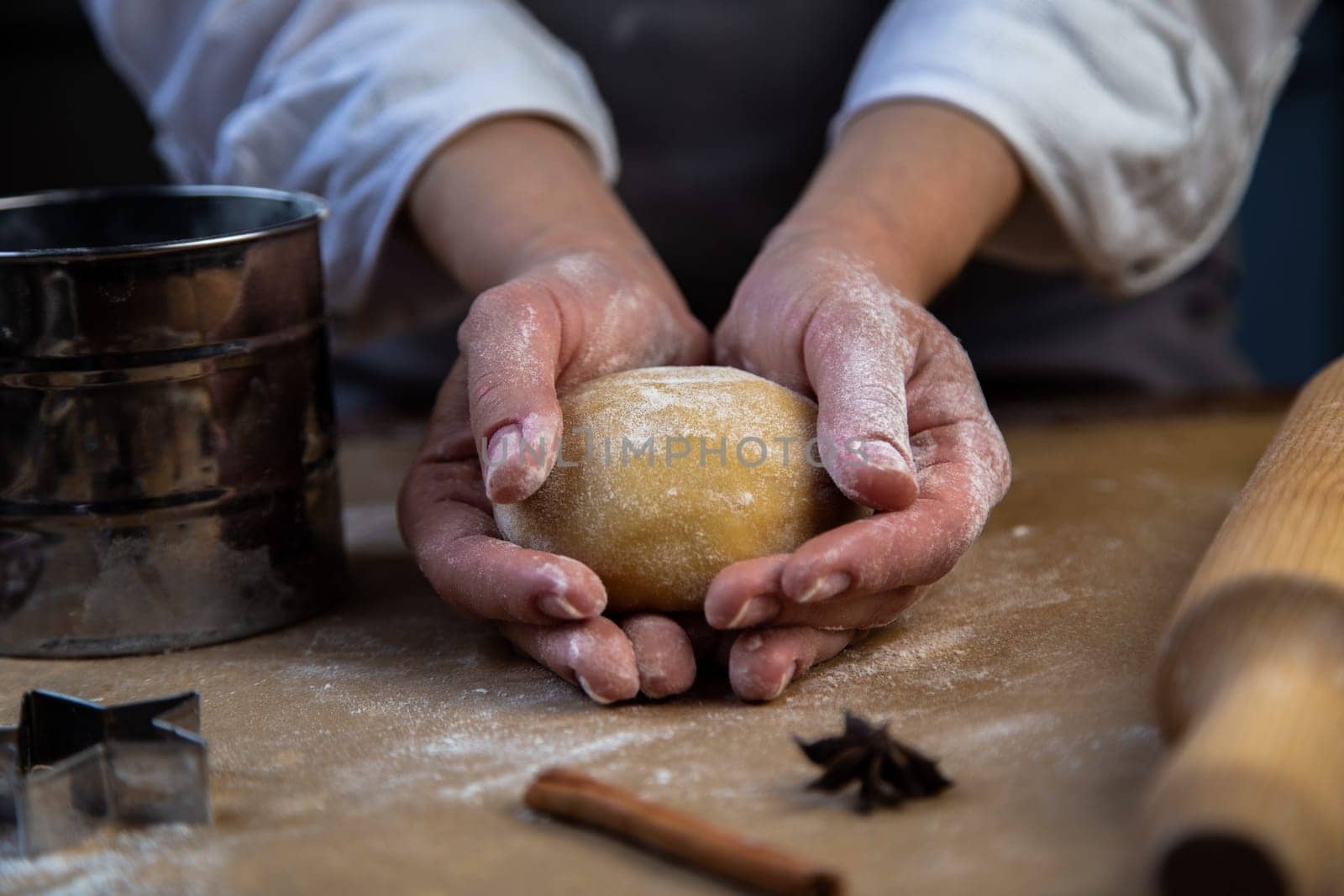 In the hands of a pastry chef, a round piece of dough, all in flour, a rolling pin, a mold with flour, cloves and cinnamon, all in dark colors, lies next to it. High quality photo
