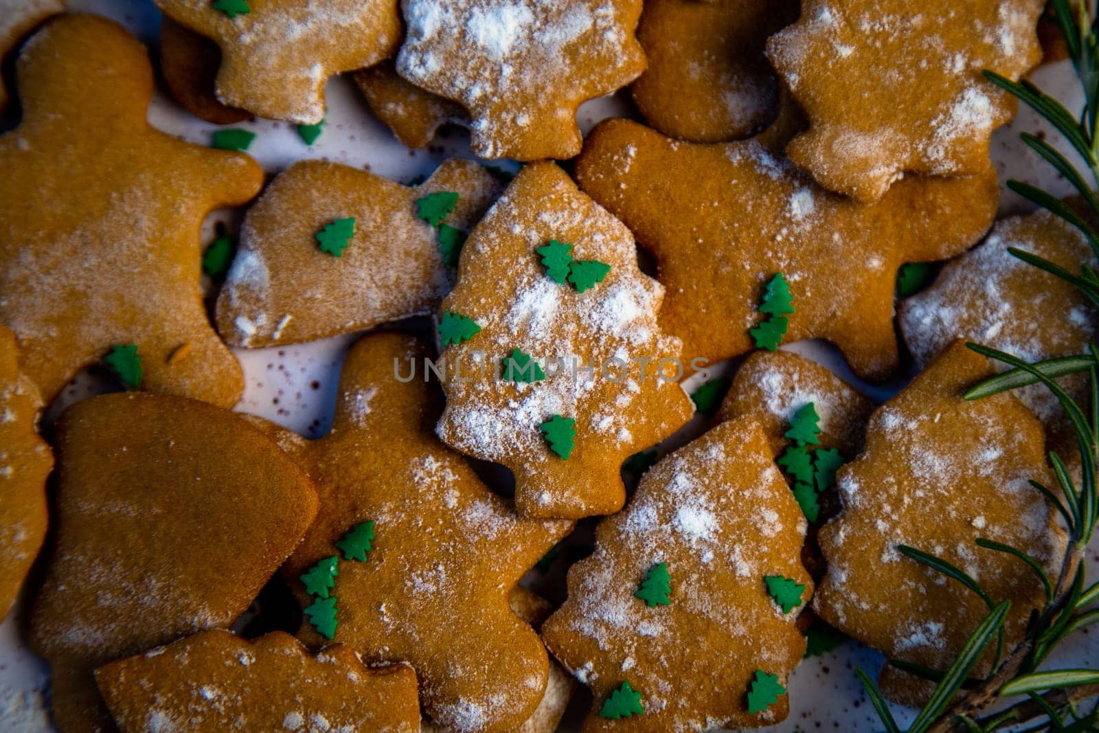 Cookies are on a plate of different shapes, in the foreground there is a cookie in the form of a Christmas tree, decorated with powdered sugar like snow and small green Christmas trees against the background of the rest of the cookies, a branch of rosemary is visible from the side. High quality photo