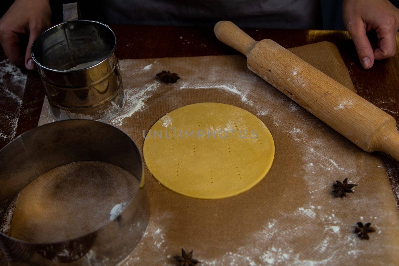 The dough, rolled out and cut into a circle, lies on the table on parchment sprinkled with flour, next to it lies a rolling pin, a cake mold and a sieve with flour. High quality photo