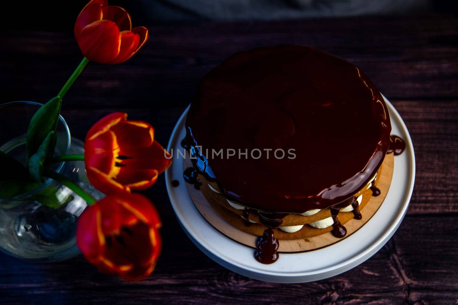 Red tulips stand in a glass vase near the cake, poured on top with chocolate icing on a dark background. Top view. High quality photo
