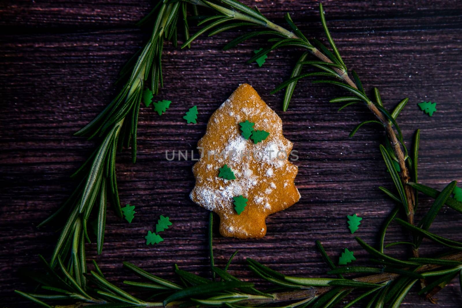 Cookies in the form of a Christmas tree, sprinkled with powdered sugar and small Christmas trees on top, lies on a dark wooden background surrounded by grained rosemary. High quality photo
