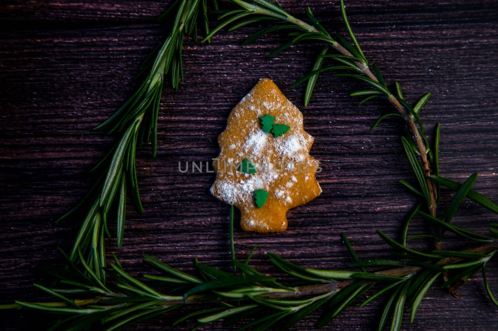 Cookies in the form of a Christmas tree, sprinkled with powdered sugar and small Christmas trees on top, lies on a dark wooden background surrounded by grained rosemary. High quality photo