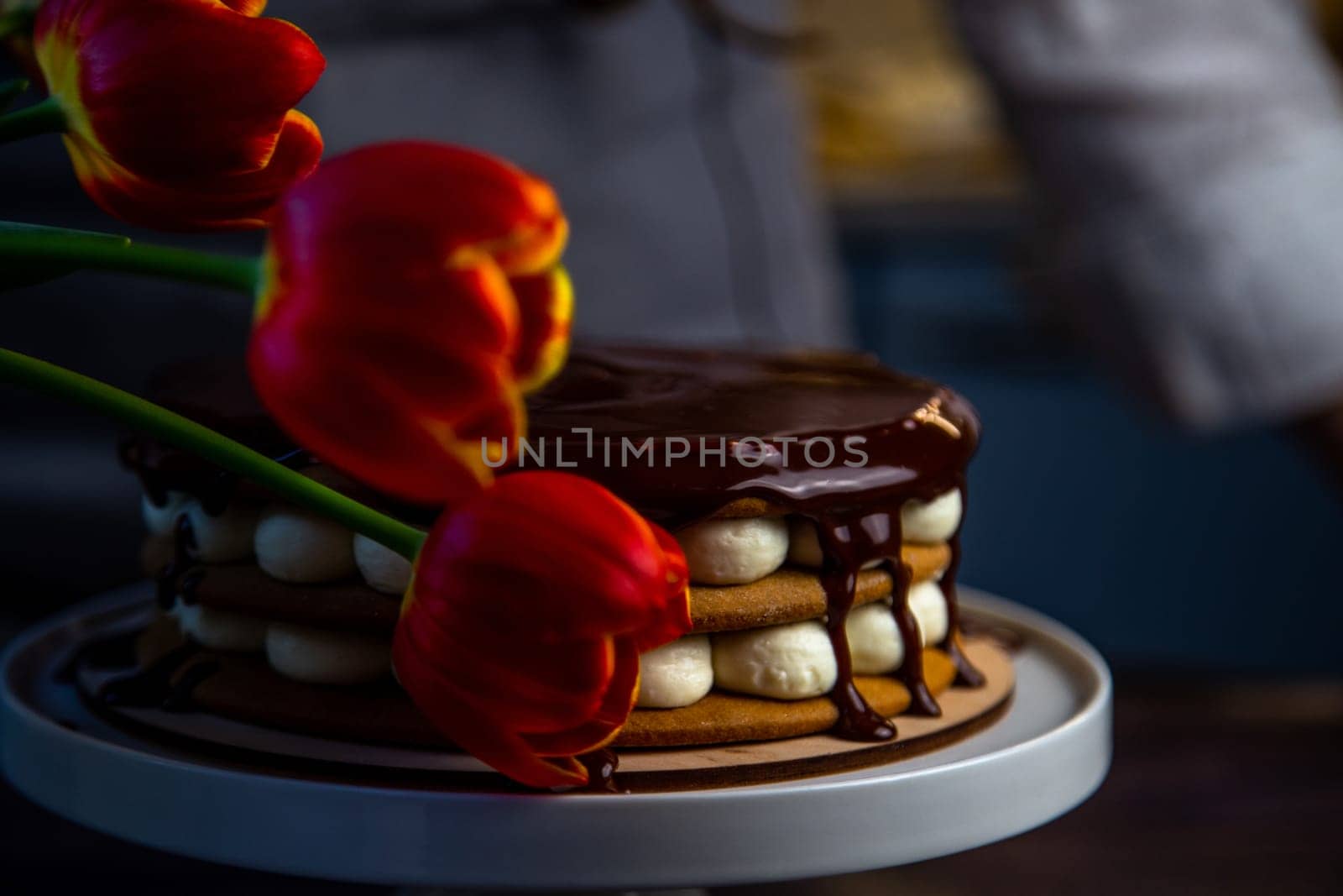 A cake with honey cakes and white cream stands on a white stand and is poured with chocolate on top, next to it are tulips, all in warm colors. High quality photo
