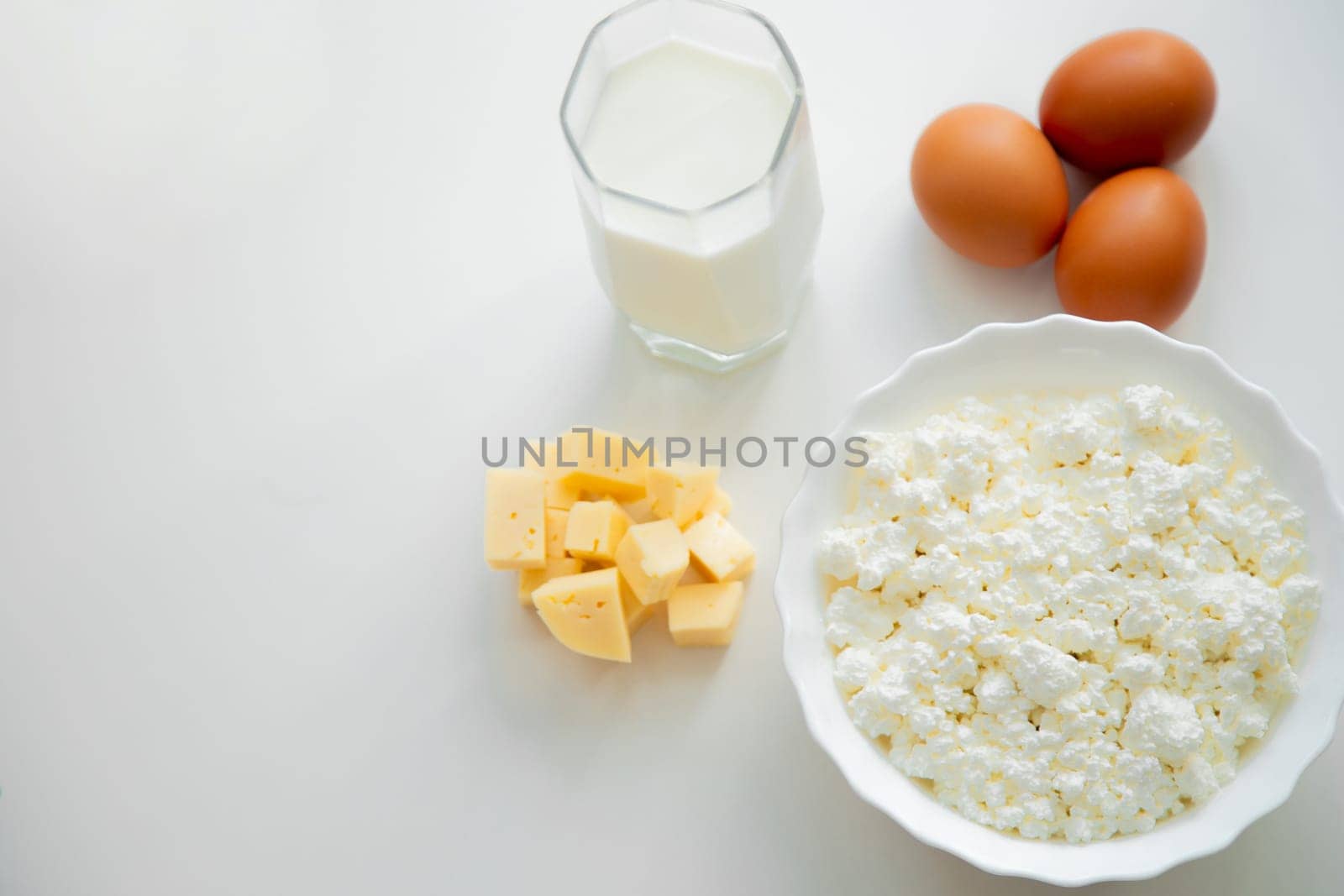 A white bowl with cottage cheese, eggs, diced cheese and a glass of milk stand on a white background. . High quality photo