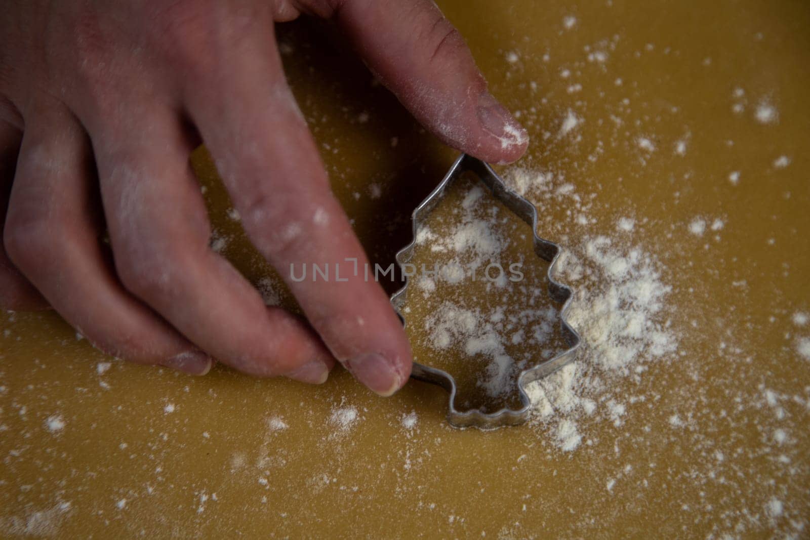 The cookie cutter in the form of a Christmas tree lies on the dough, which is sprinkled with flour like snow, and the pastry chef's hand holds the mold. High quality photo