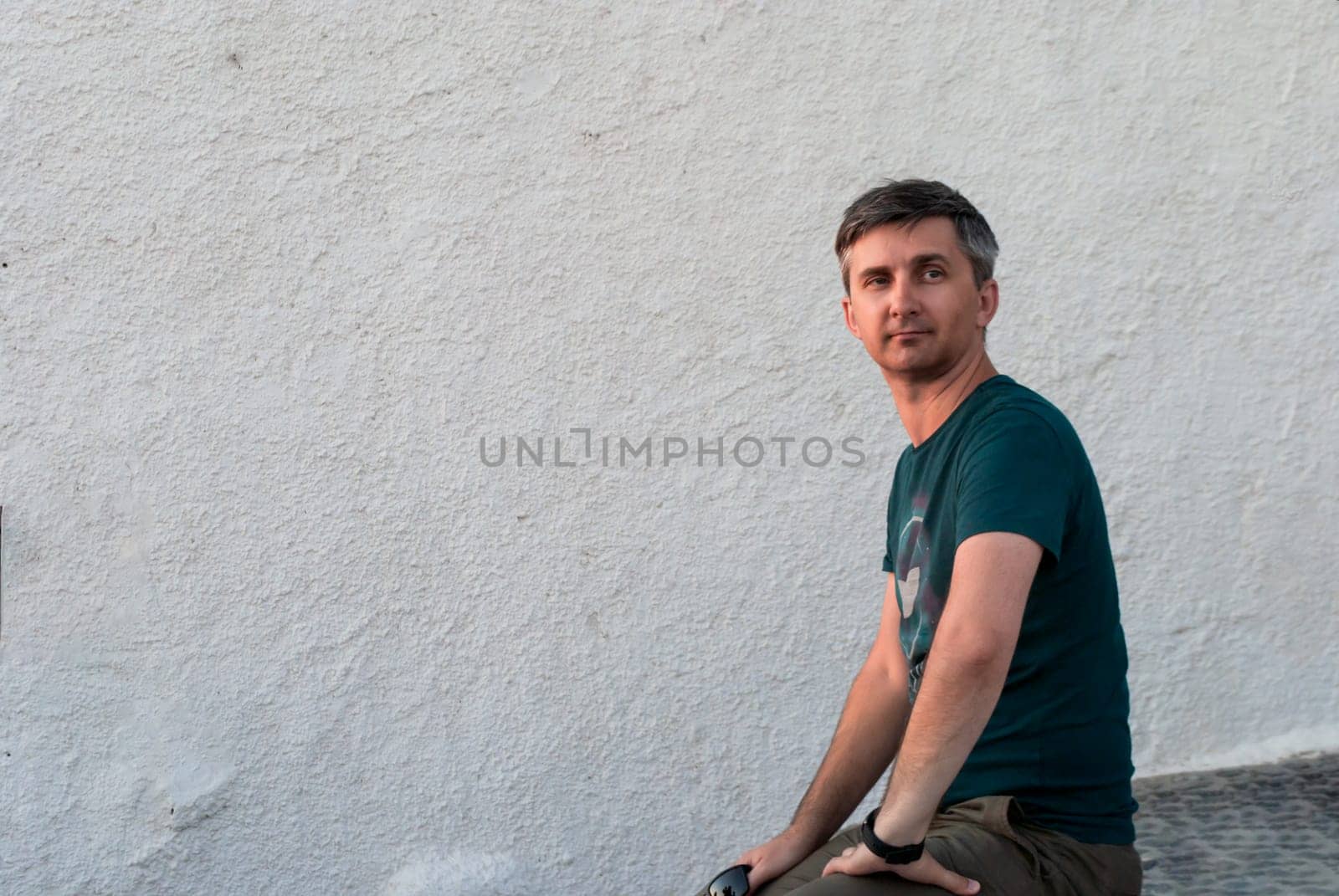 A middle-aged man sits against a white wall in summer clothes
