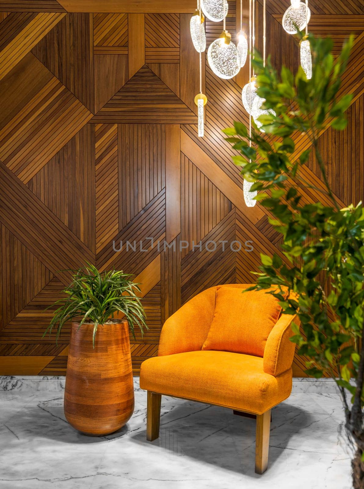 Modern orange armchair, tall wooden planter, and contemporary glass chandelier, in a hall with decorated wood cladding wall, and white marble floor, framed by green bushes