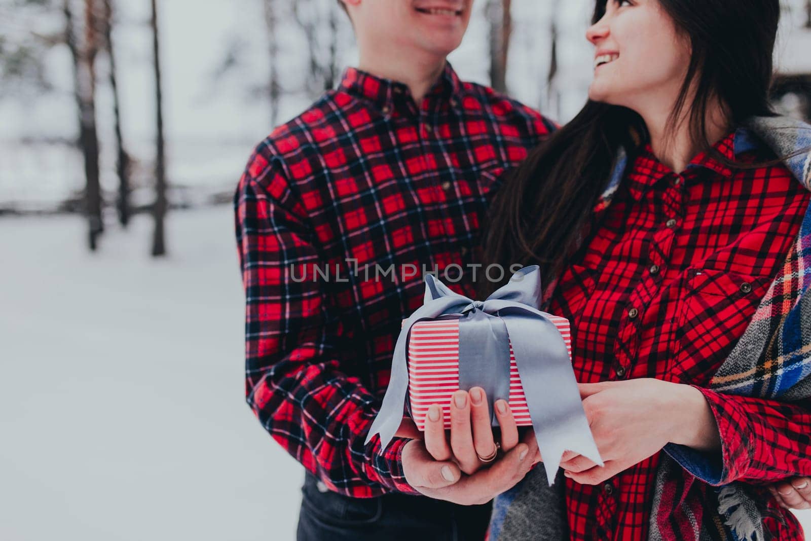 Boyfriend giving a gift box present with ribbon outdoor due to Saint Valentine day to her girlfriend. Snowy forest, trees with snow. Two are dressed in red clothes. Beautiful couple love each other