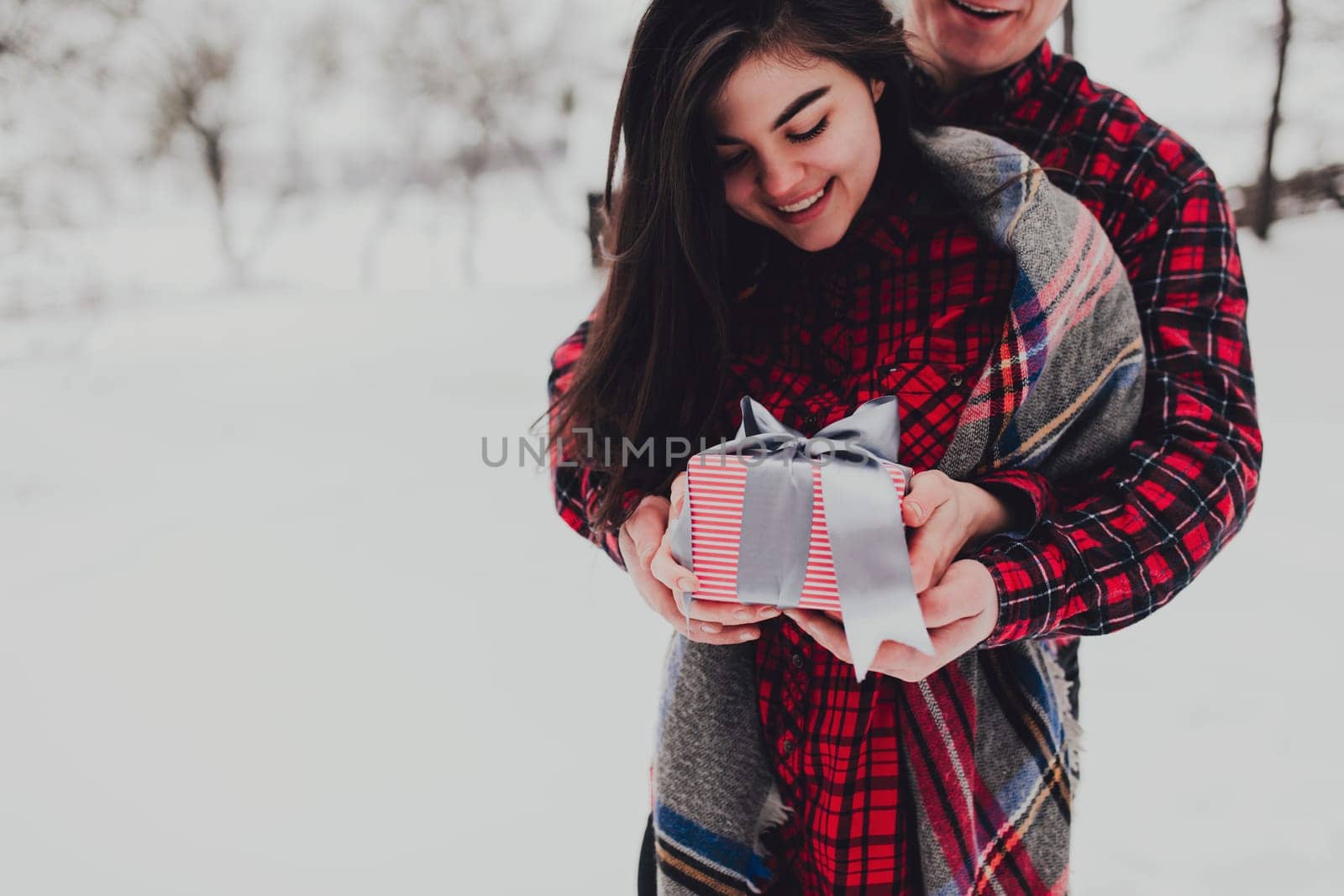 Boyfriend giving a gift box present with ribbon outdoor due to Saint Valentine day to her girlfriend. Snowy forest, trees with snow. Two are dressed in red clothes. Beautiful couple love each other