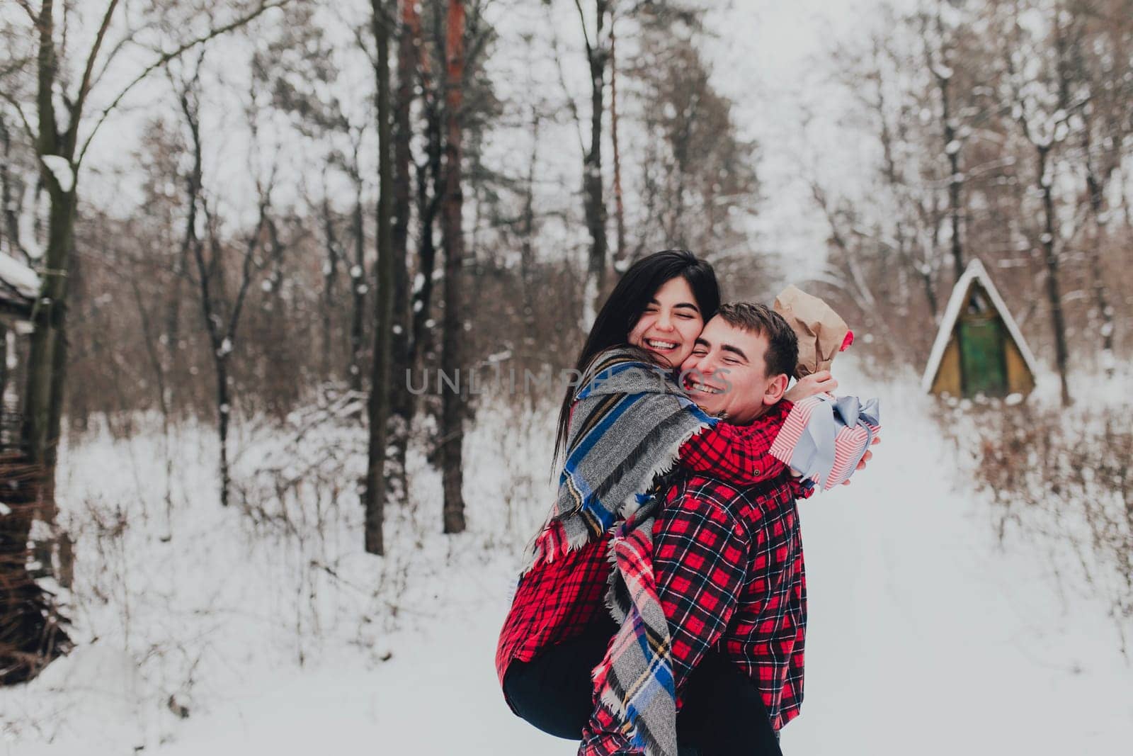 Boyfriend giving a gift box present with ribbon outdoor due to Saint Valentine day to her girlfriend. Snowy forest, trees with snow. Two are dressed in red clothes. Beautiful couple love each other