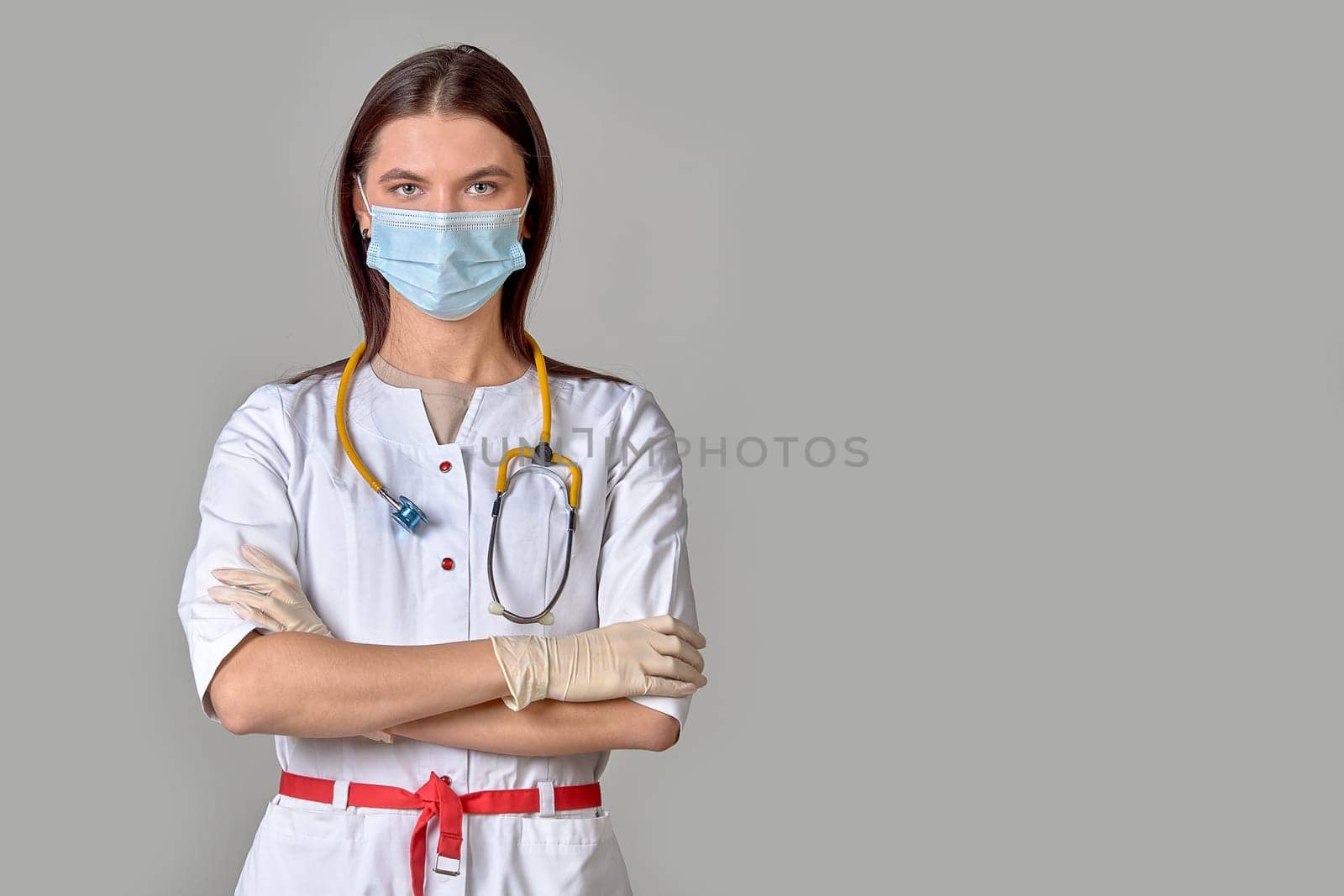 A young female doctor in a white coat with a stethoscope and a medical mask on a gray isolated background. Copy space. High quality photo