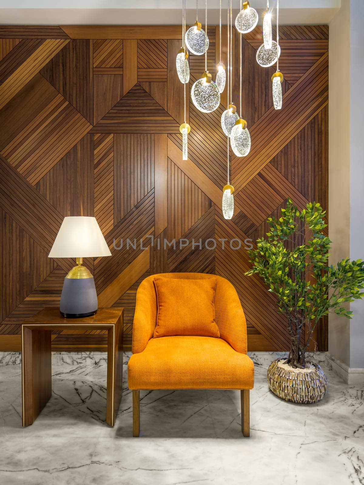 Modern orange armchair, lamp shade on small wooden table, planter with green bushes, and contemporary tall glass chandelier, in a hall with decorated wood cladding wall, and white marble floor