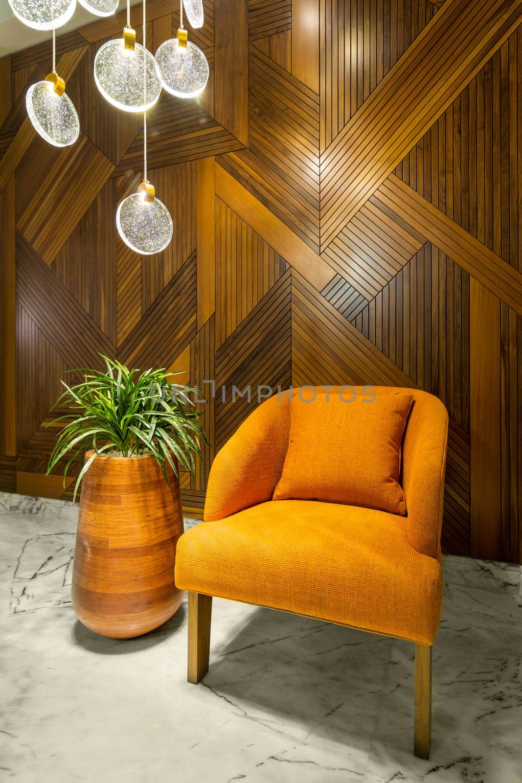 Modern orange armchair, tall wooden planter with green bushes, and contemporary glass chandelier, in a hall with decorated wood cladding wall, and white marble floor