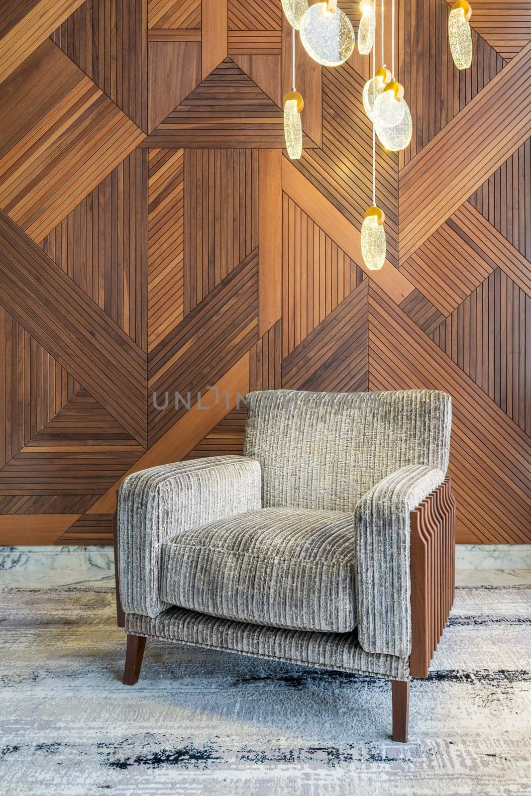 Modern grey armchair, and contemporary tall glass chandelier, in a hall with decorated wood cladding wall, and grey modern carpet