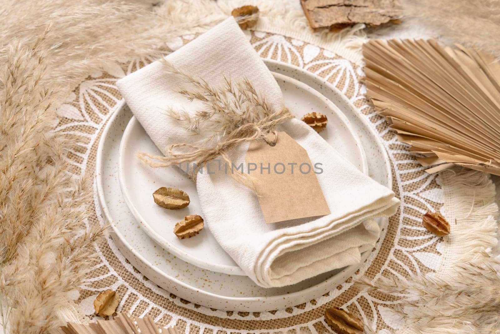 Wedding table place with blank gift tag attached to a napkin on bohemian placemat top view. Boho wedding label mockup Tropical beige template with dried plants, palm leaves and pampas grass, flat lay