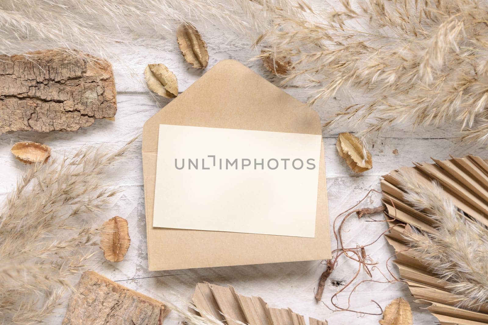 Blank card and envelope on white wooden table near dried plants, palm leaves and pampas grass top view. Boho scene with horizontal paper card flat lay. Greeting or Invitation card mockup, place for text