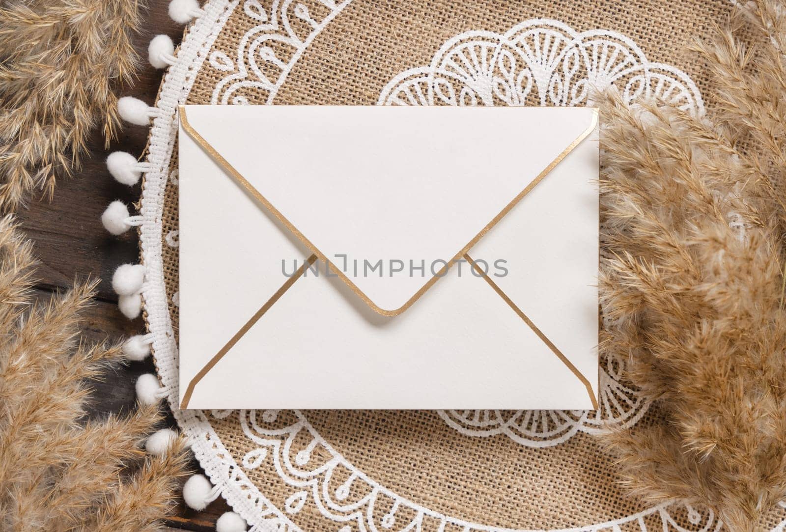 Blank envelope on bohemian jute placemat near pampas grass top view. Boho rustic scene with greeting or Invitation envelope mockup, flat lay.