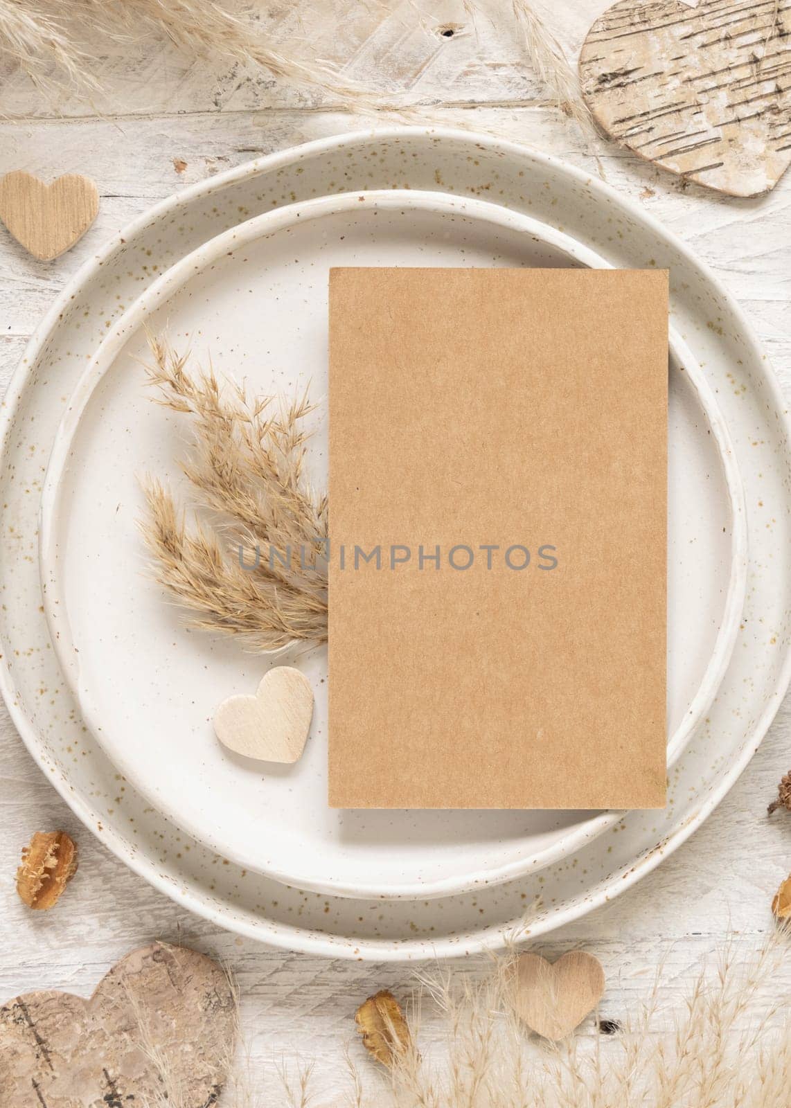 Wedding table place with blank card near dried plants, palm leaves, pampas grass and hearts top view. Boho scene with vertical kraft paper card flat lay. Greeting, Invitation or valentines mockup