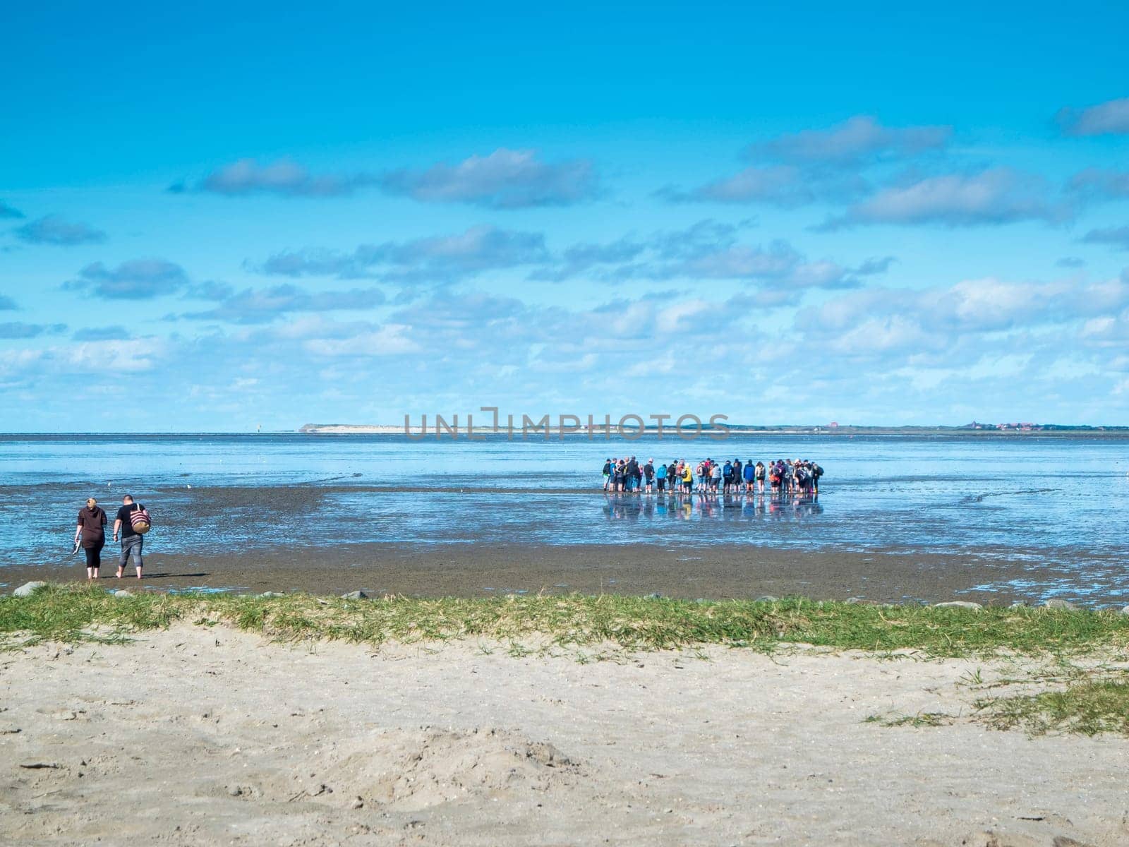Landscape view of the North German Wadden Sea at low tide with a guided group of mudflat hikers in a blue sky with clouds.