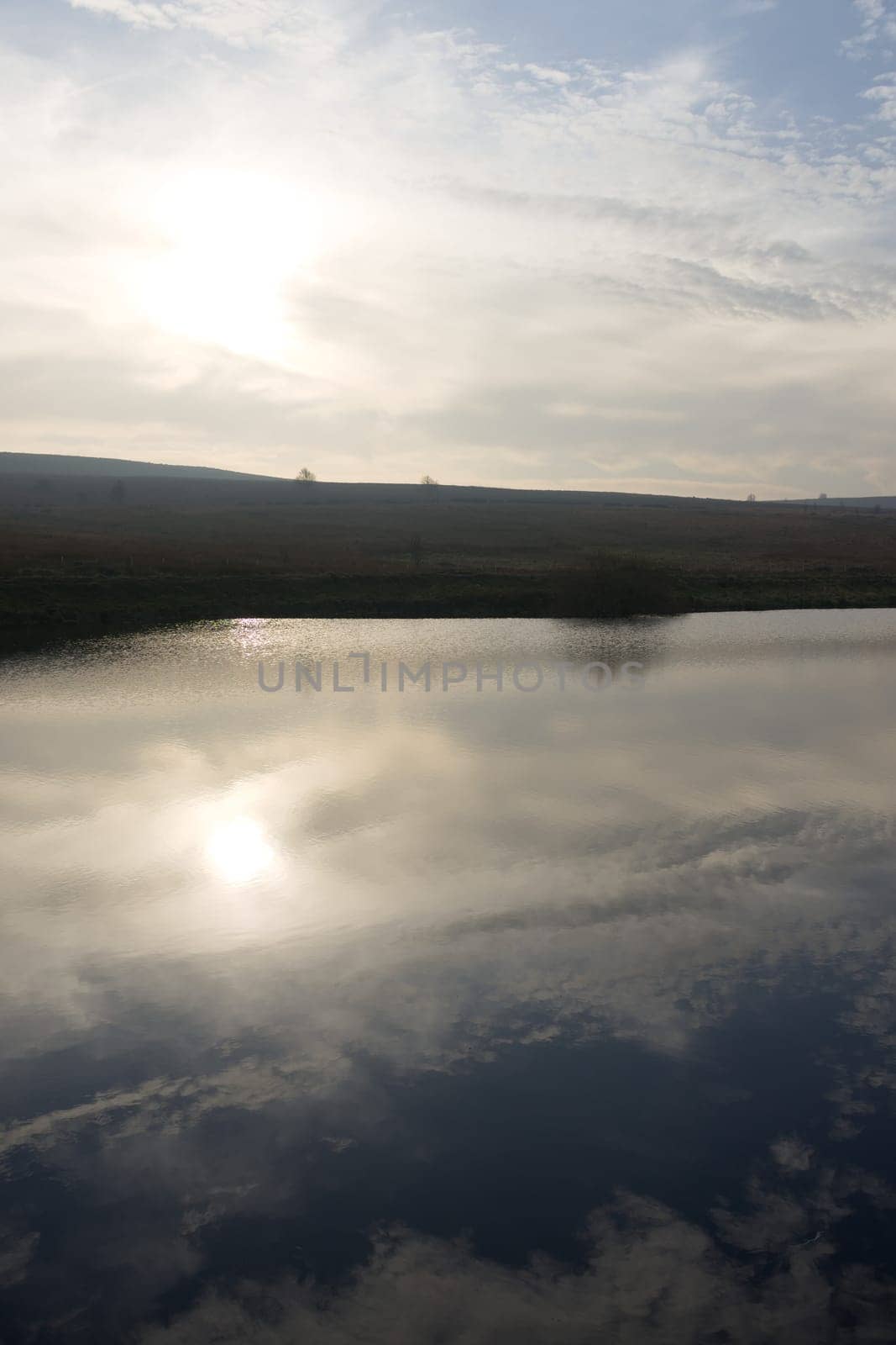 Vertical image of blue cloudy sky reflected in redmires top reservoir. Looking into the sun over heather covered peak district moor. Winter season.
