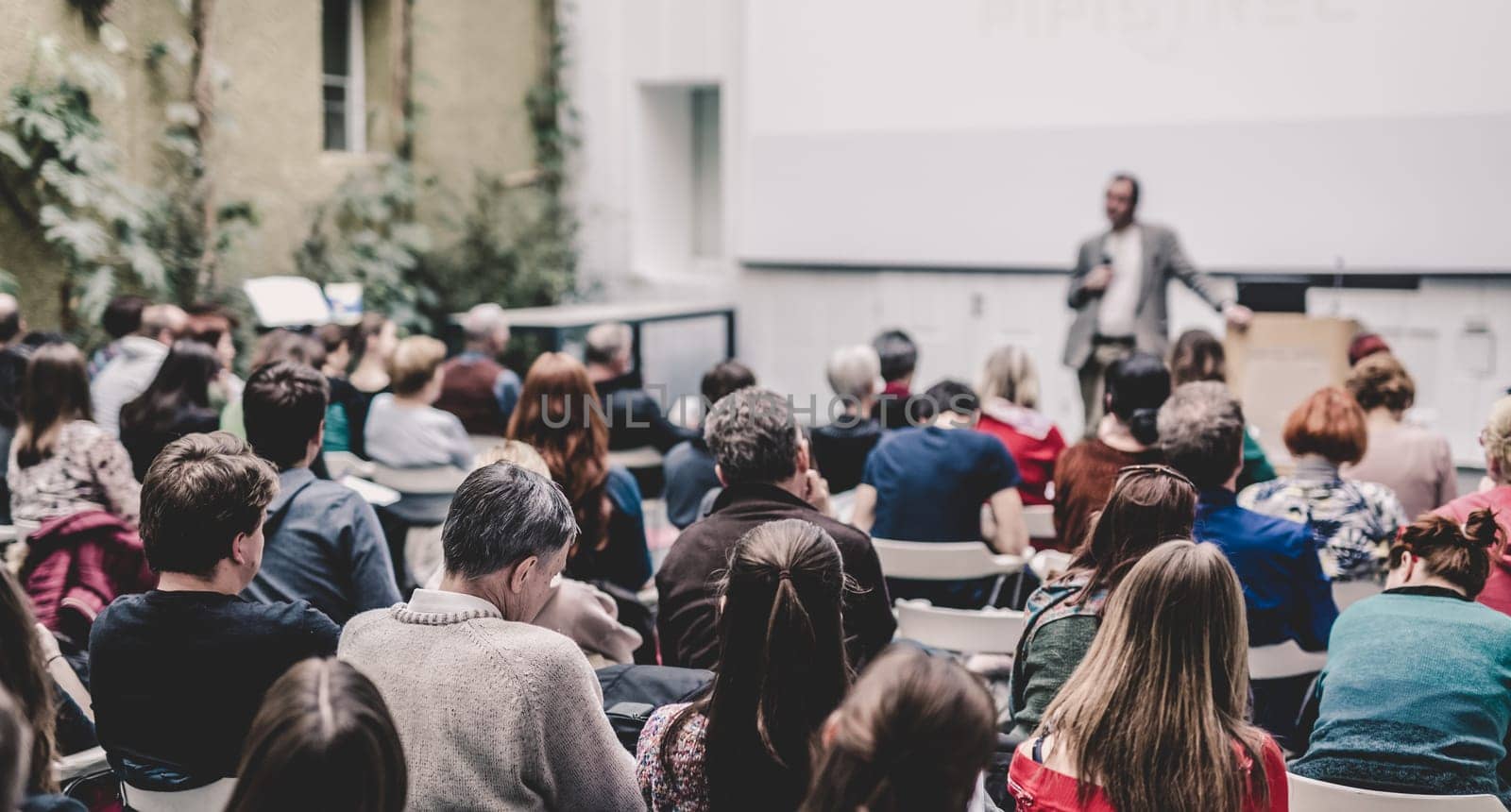 Male speaker giving presentation in lecture hall at university workshop. Audience in conference hall. Rear view of unrecognized participant. Scientific conference event. Copy space on whitescreen.
