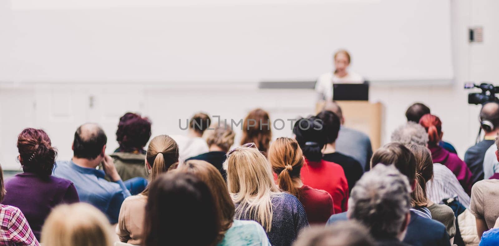 Business and entrepreneurship symposium. Female speaker giving a talk at business meeting. Audience in conference hall. Rear view of unrecognized participant in audience. Copy space on whitescreen.