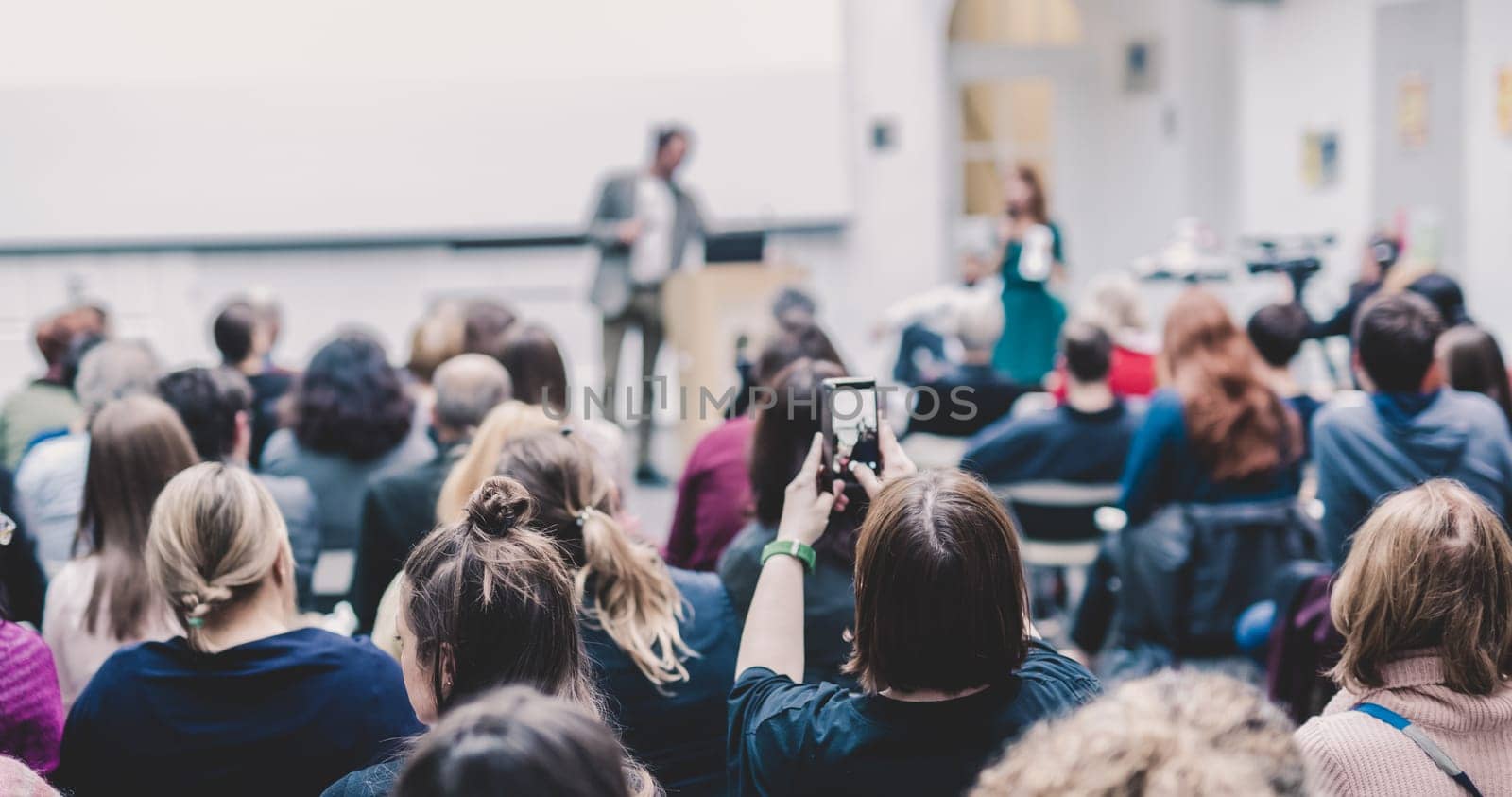 Male speaker giving presentation in lecture hall at university workshop. Audience in conference hall. Rear view of unrecognized participant in audience. Scientific conference event.