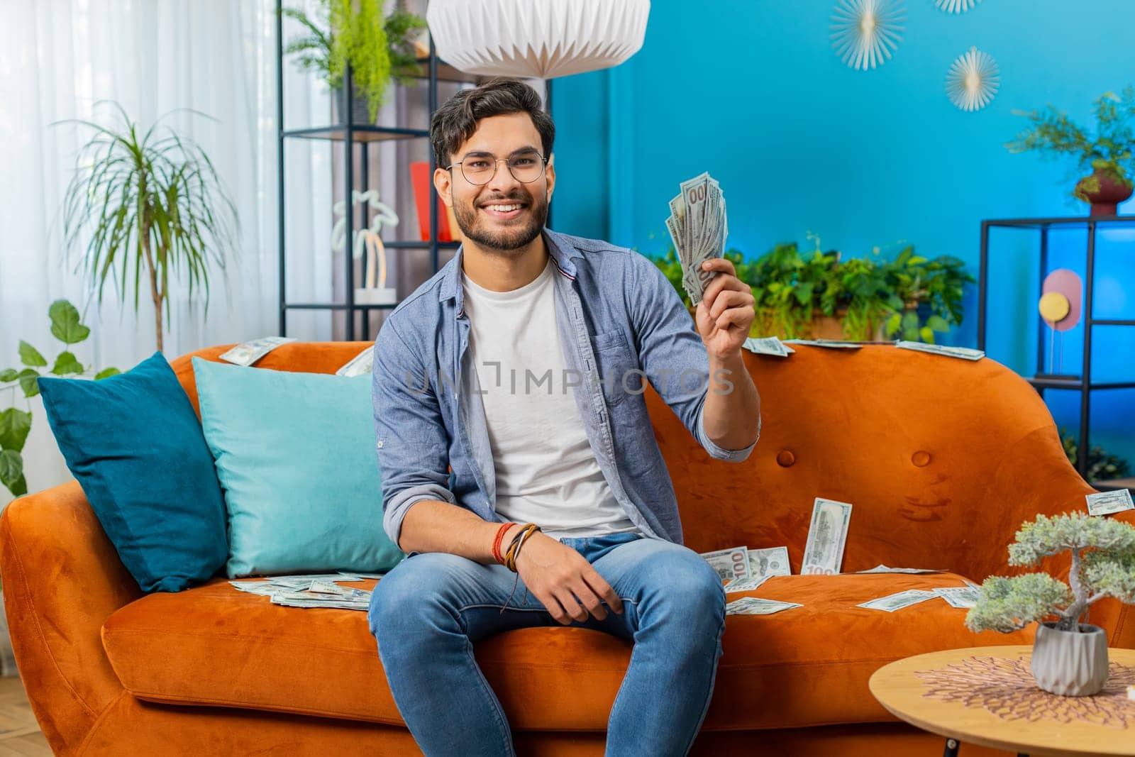 Successful happy rich winner Indian Arabian man counting money cheering up with high profits, lottery game win, satisfied of wealth income earnings salary, on home couch full of dollar cash banknotes